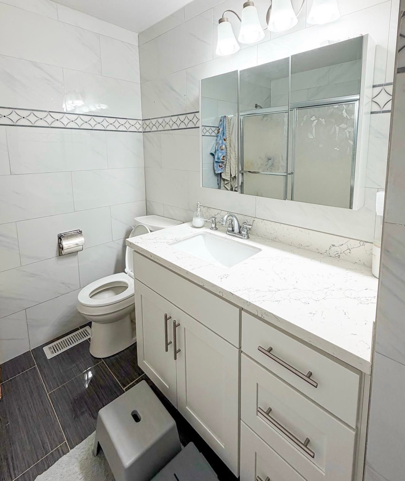 White bathroom with vanity, toilet, and shower. Gray and white tile, mirror, and white step stool.