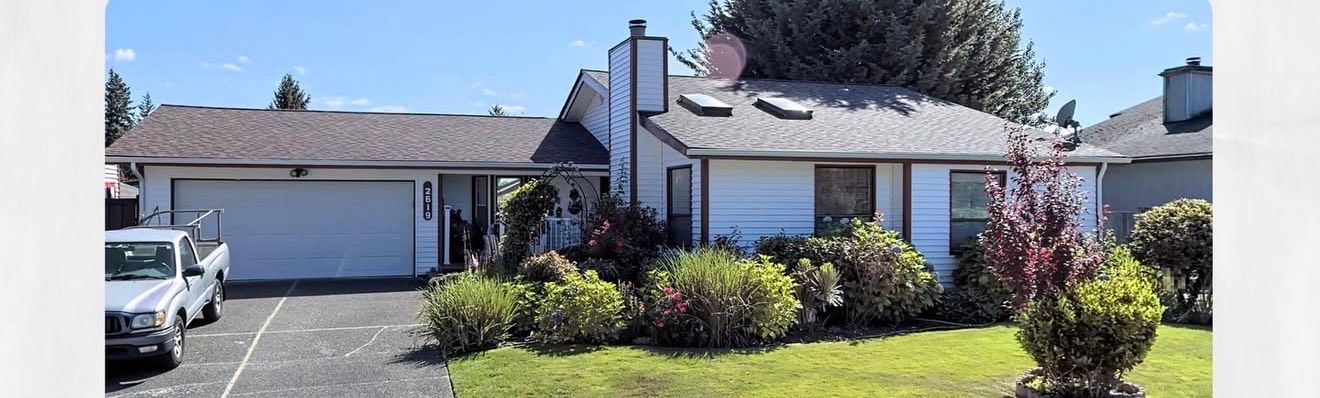 A white house with a gray roof and a truck parked in the driveway. The house is surrounded by green bushes.