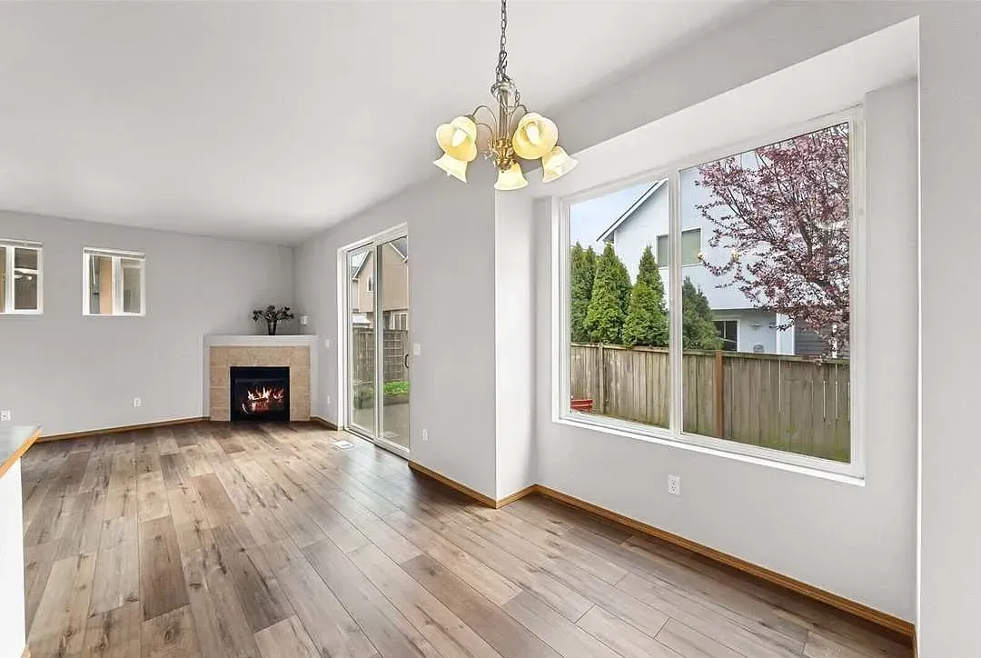 Spacious room with fireplace, sliding door, and large window overlooking a yard; wooden floors and chandelier.