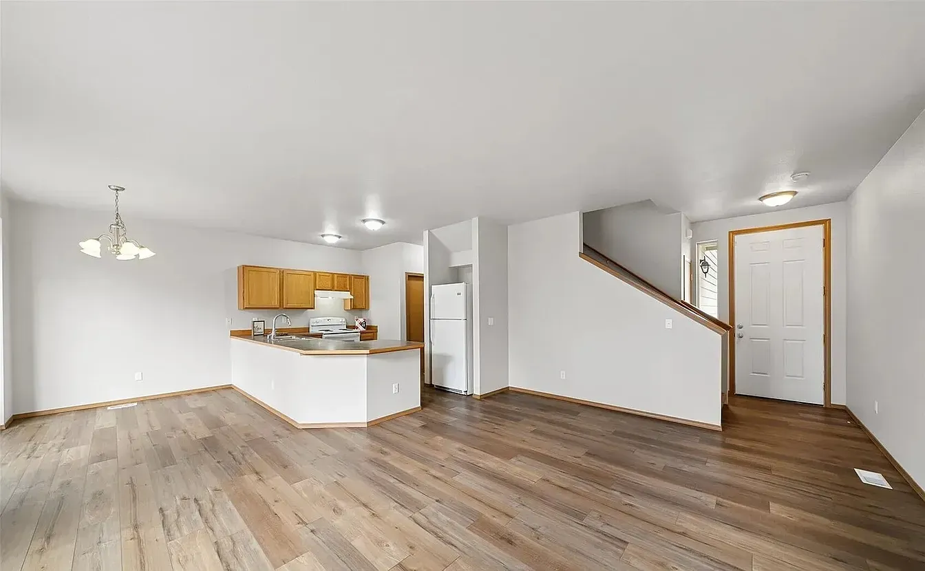 Empty, open-concept living space with hardwood floors, kitchen, staircase, and white walls.