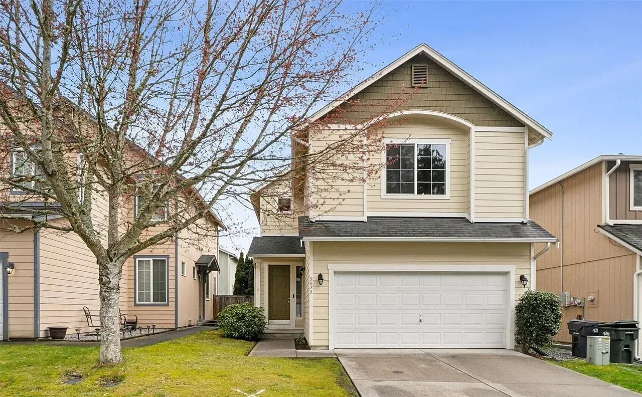 Two-story house with a beige exterior, a garage, and a bare tree in the front yard.