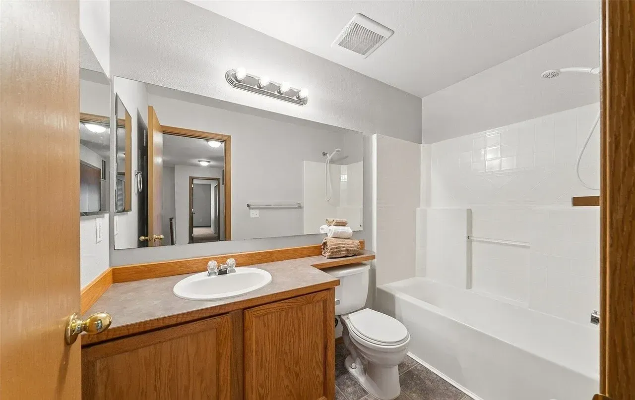 Bathroom with a tub, toilet, vanity, and large mirror. White and gray tones, wooden cabinets, and a door on the left.