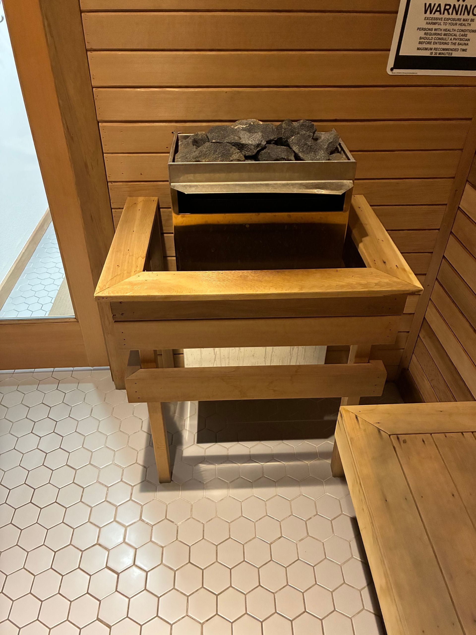 A sauna interior with a wooden heater, rocks, and a bench. Light wood walls and a tiled floor.