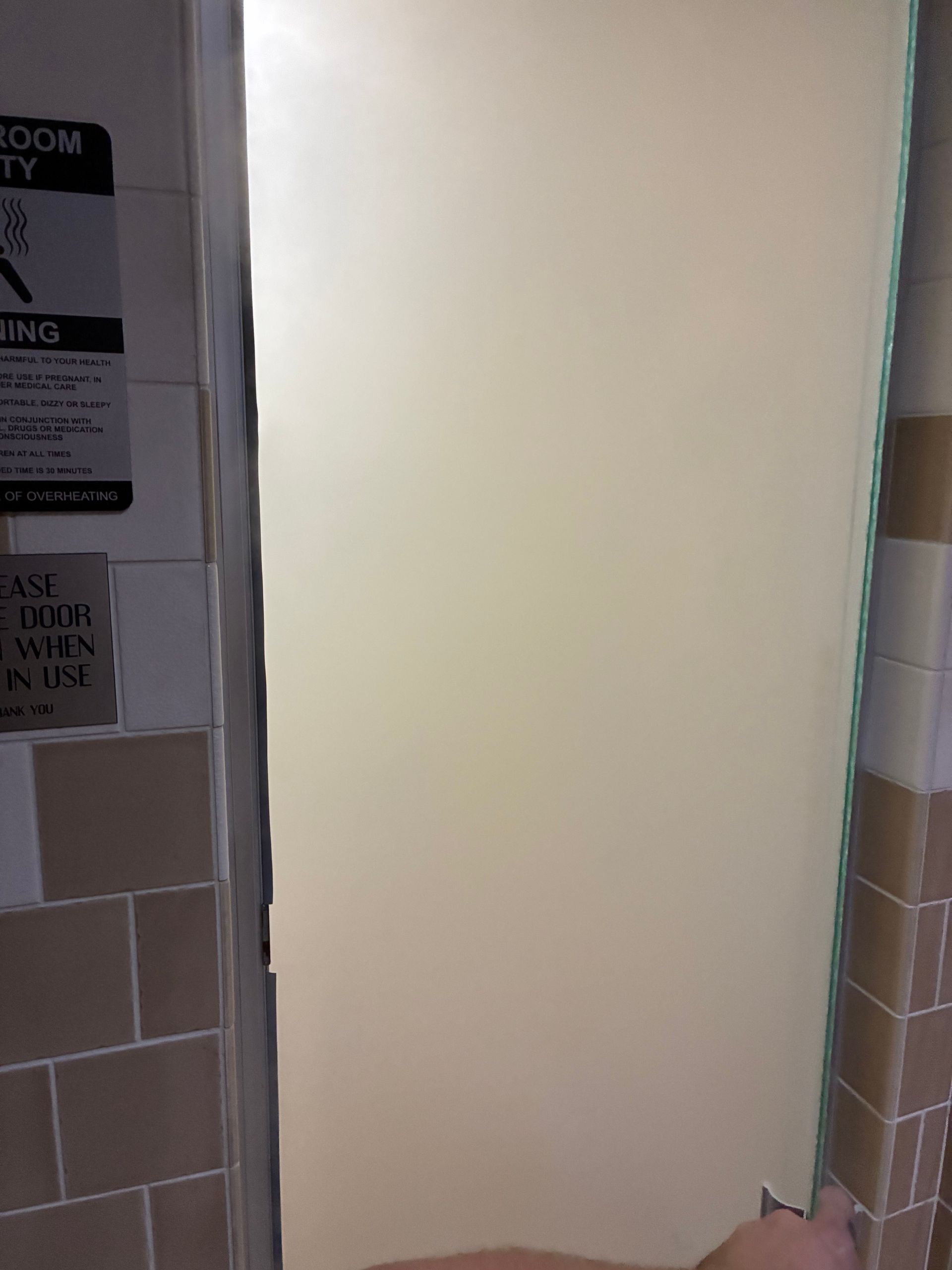 Bathroom stall doorway, white wall, tan and brown tiled walls. Sign on left.