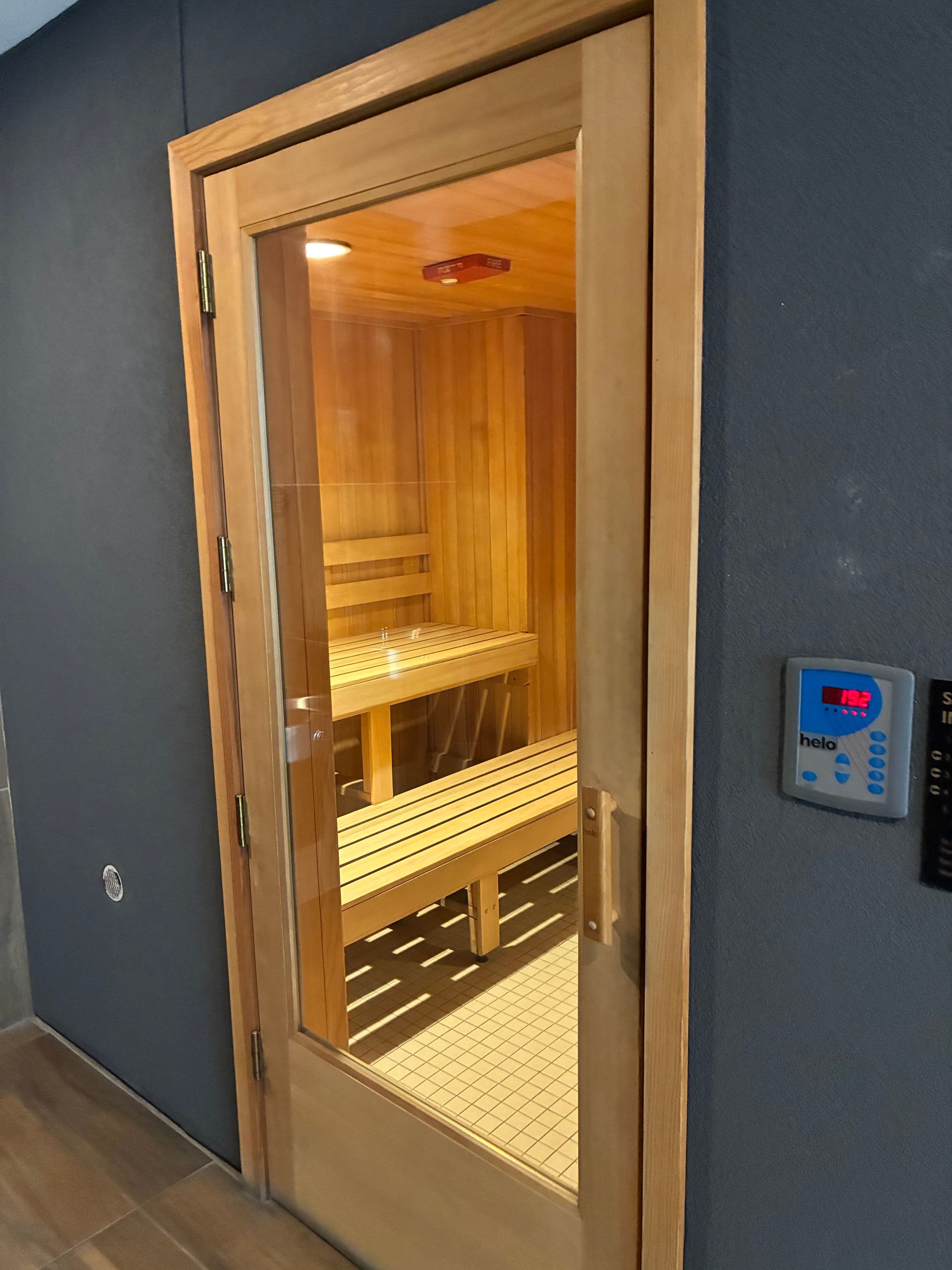 Wooden sauna with glass door, interior view. Bench seating, control panel on adjacent wall.