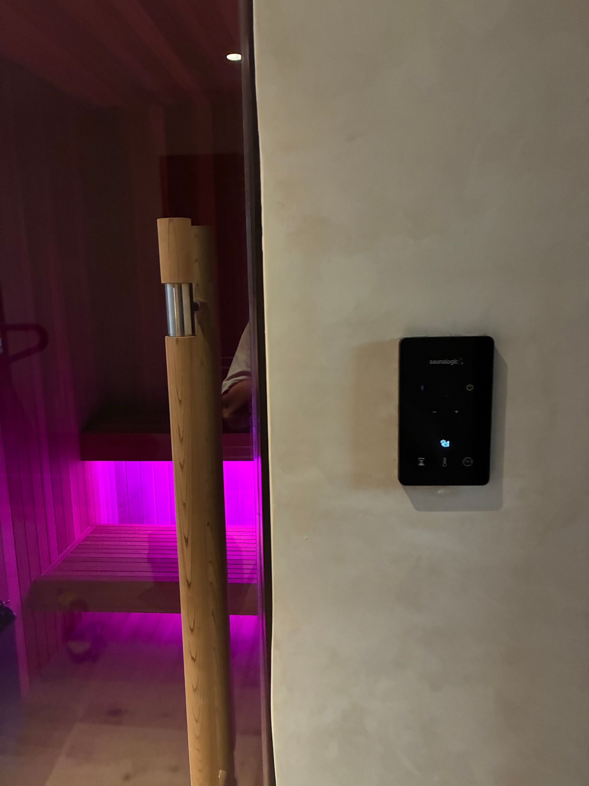 Sauna interior with control panel on wall. Pink light under benches; wooden door handle.
