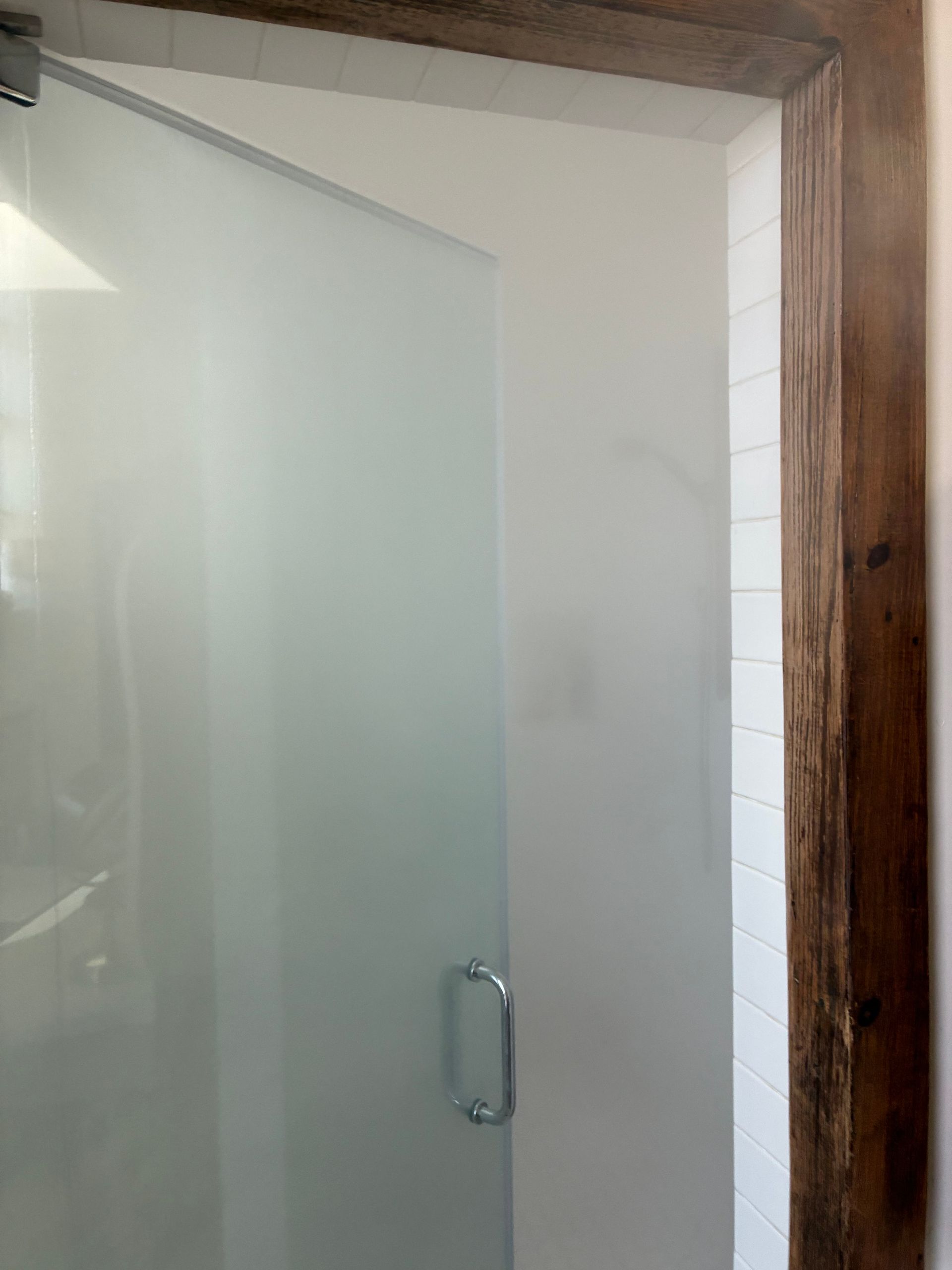 Frosted glass shower door with chrome handle, set in a wood frame, adjacent to white tiled wall.