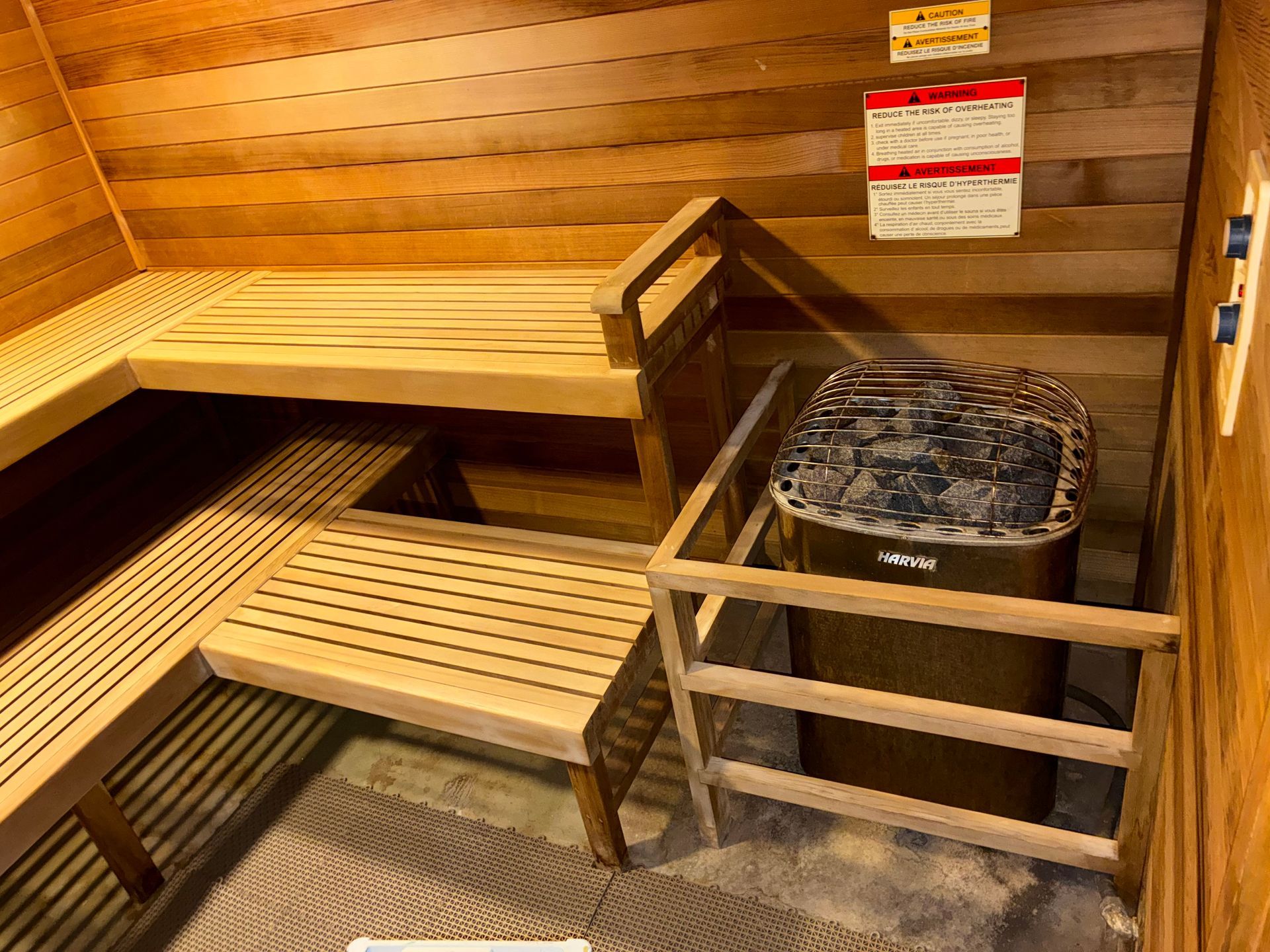Interior of a wooden sauna with tiered benches and a heater enclosed by a wooden fence; a control panel and signs are on the right.