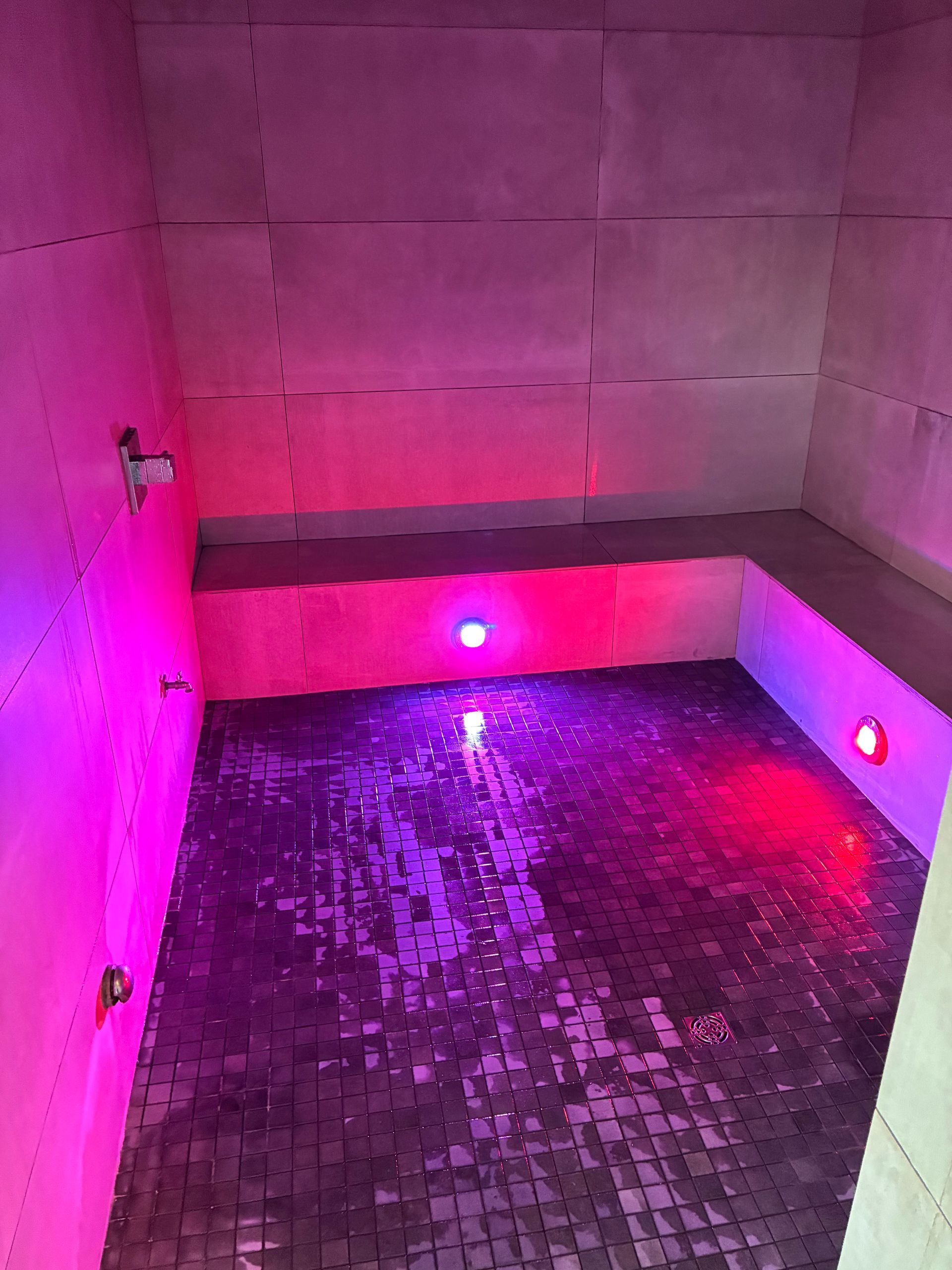 A small steam room with pink and purple lights, tiled walls and floor, and a built-in bench.