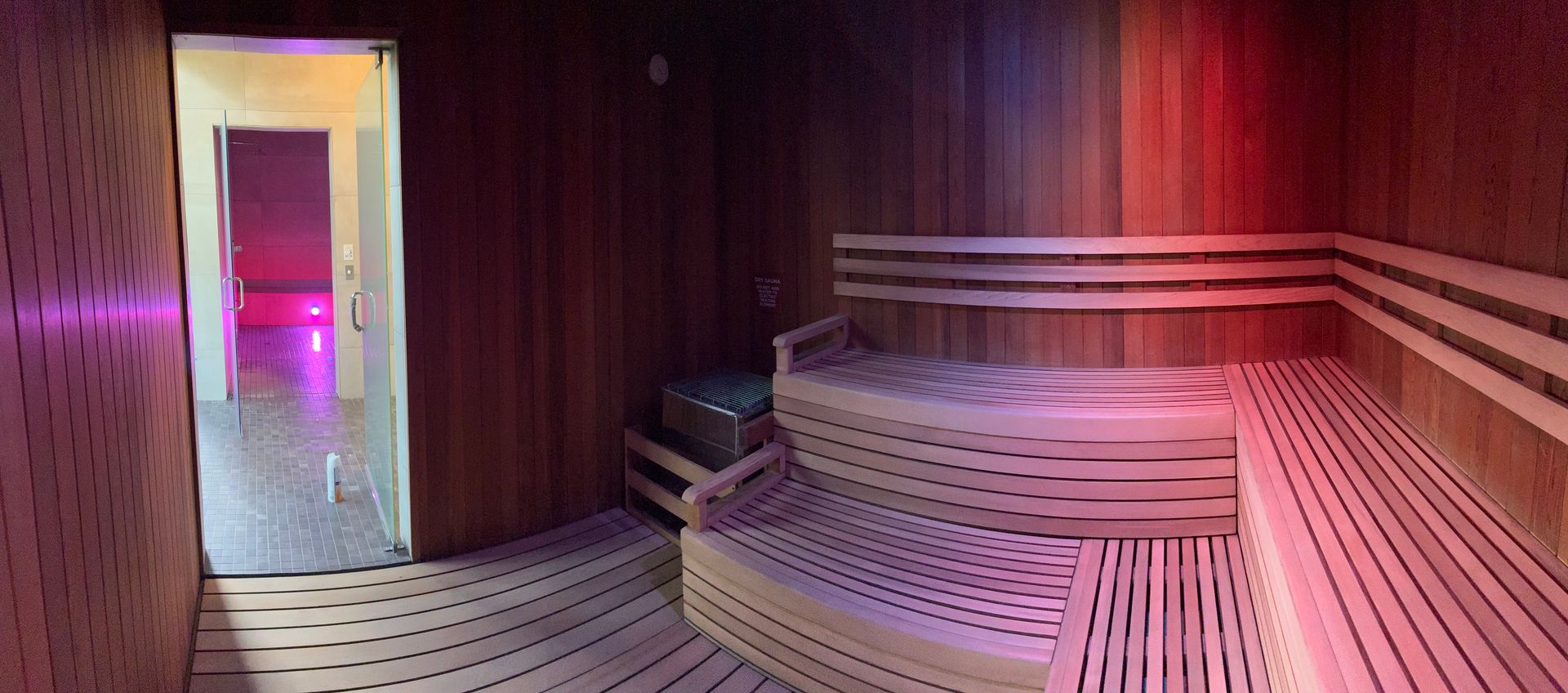 Interior of a wooden sauna with tiered seating, glass door to a hallway with colorful lighting.