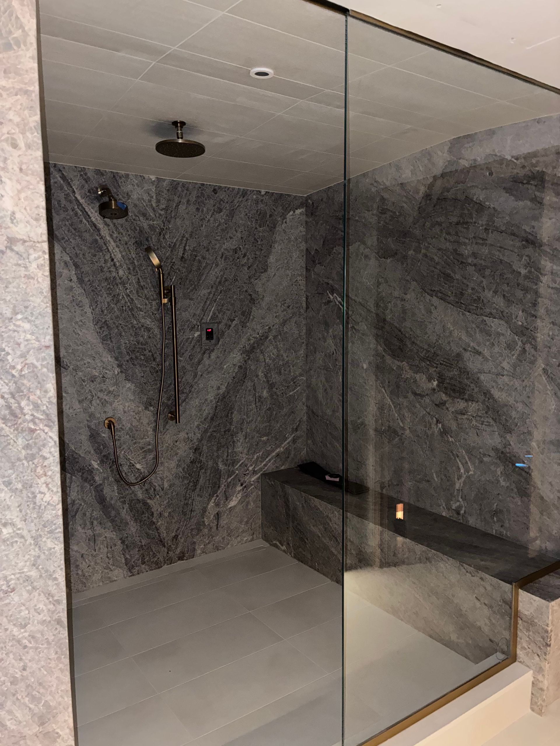Modern shower with dark gray marbled walls, glass door, and bench.