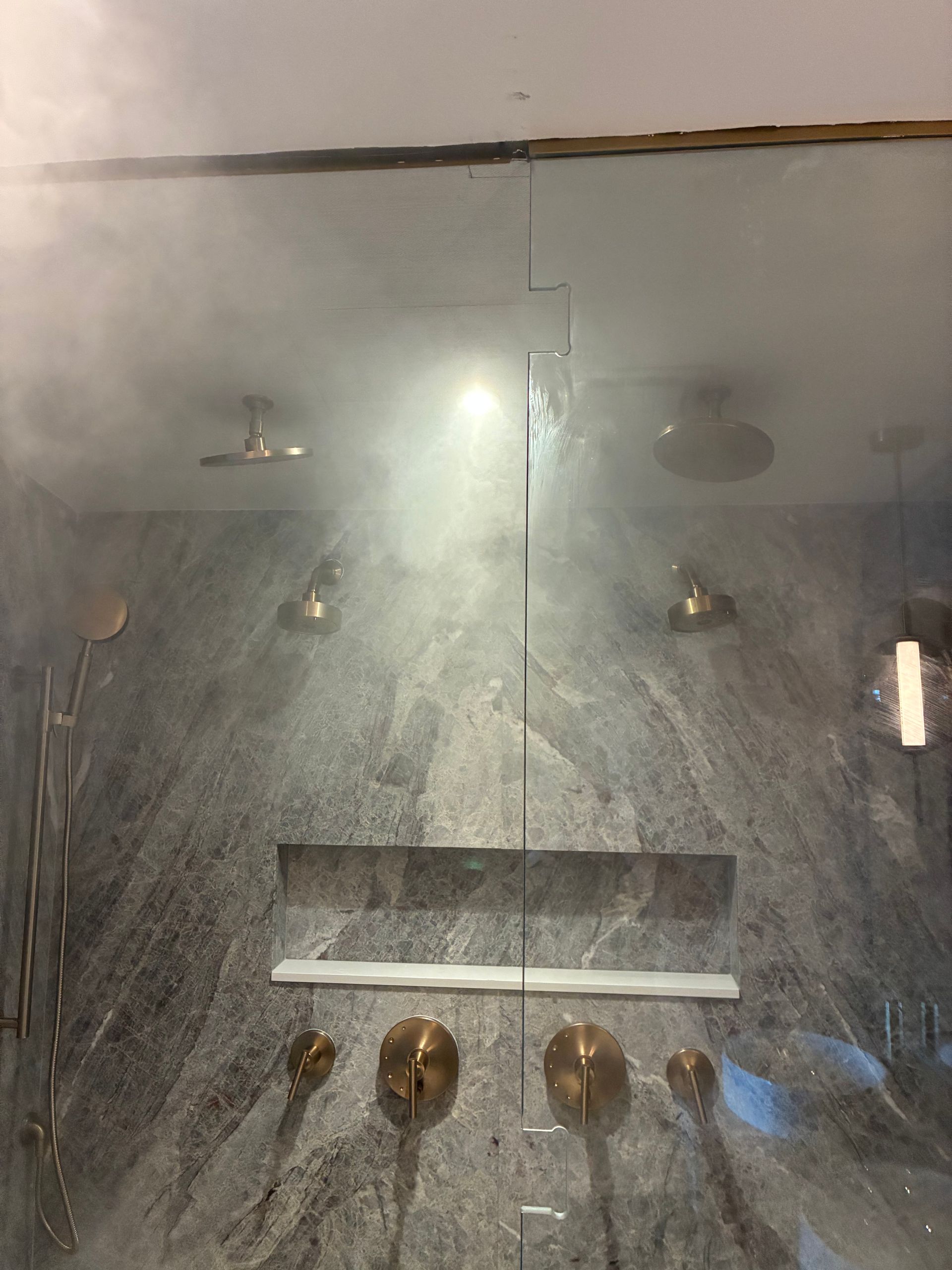 Steamy shower with multiple gold showerheads on a gray stone wall; light shines through the mist.