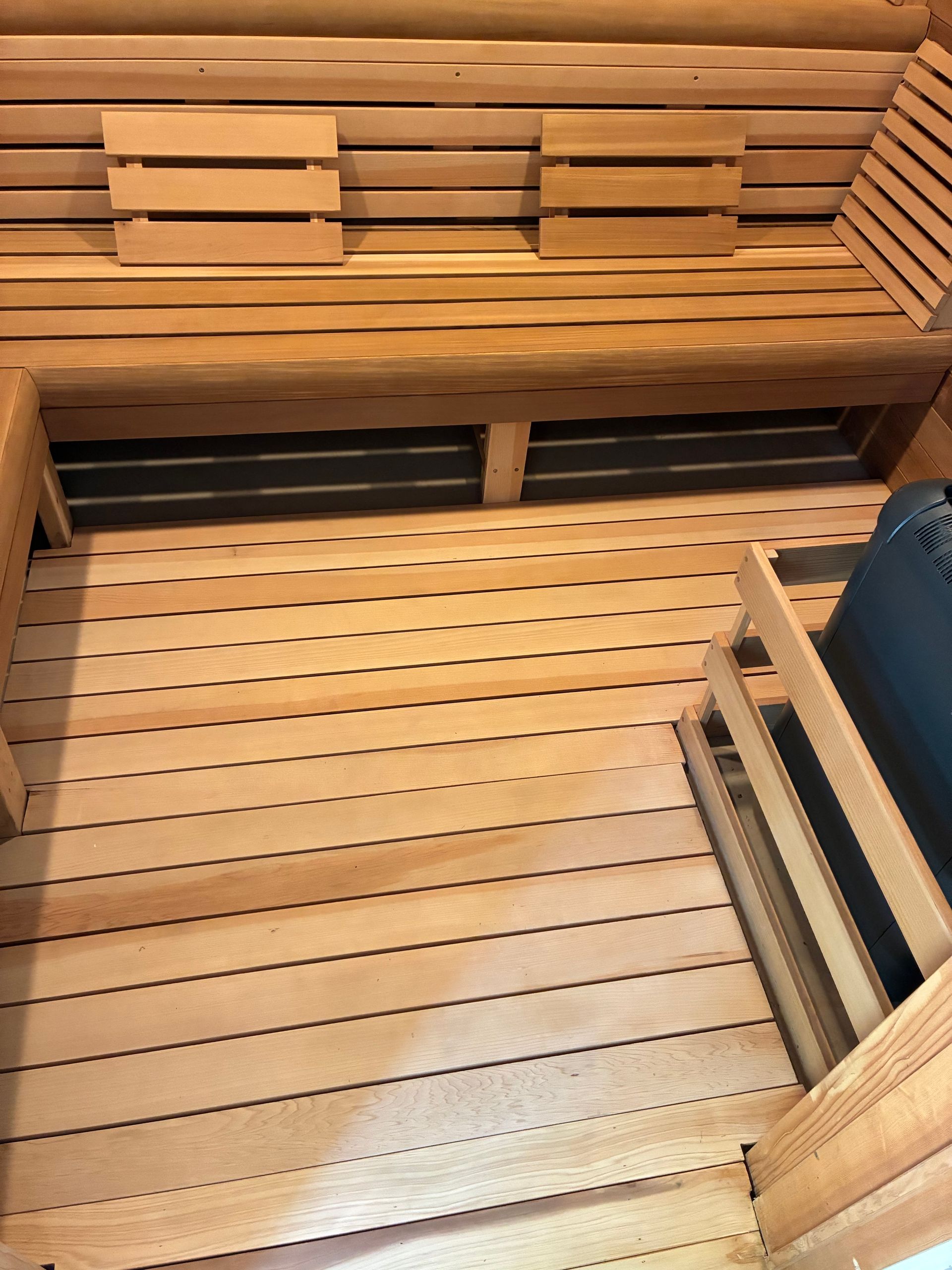 Interior of a wooden sauna with benches and slatted floor. A dark heater is in the corner.