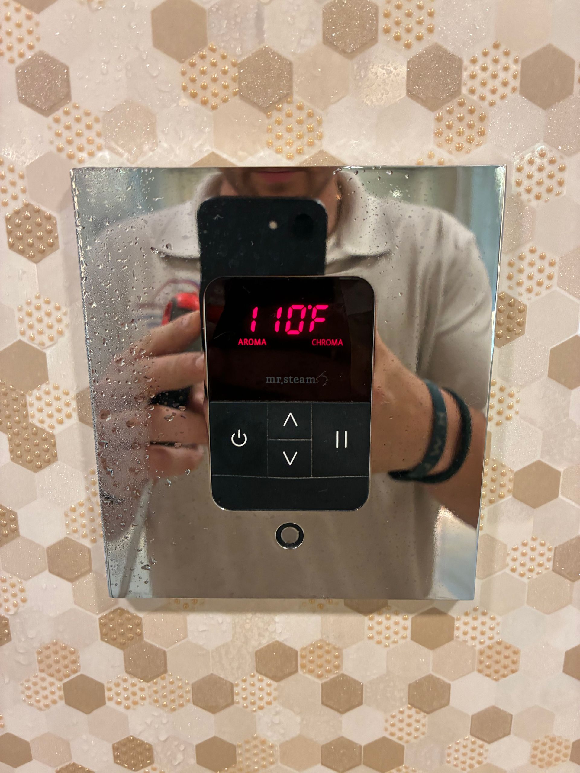 A digital shower controller displays 110°F. A person in a tan shirt is reflected in the mirror.