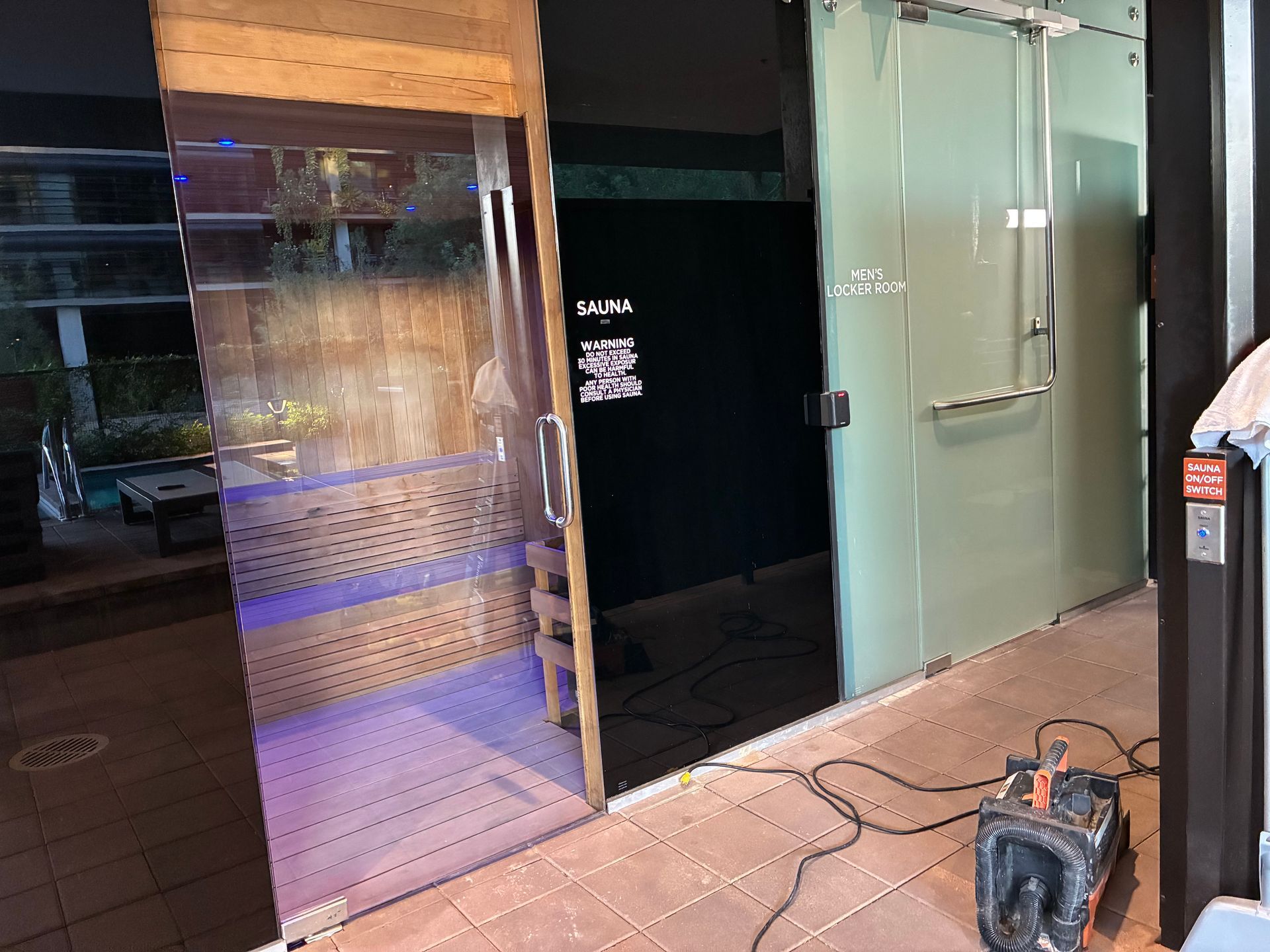 Sauna with glass doors, and wooden interior. Electrical equipment and a partially visible pool in the setting.