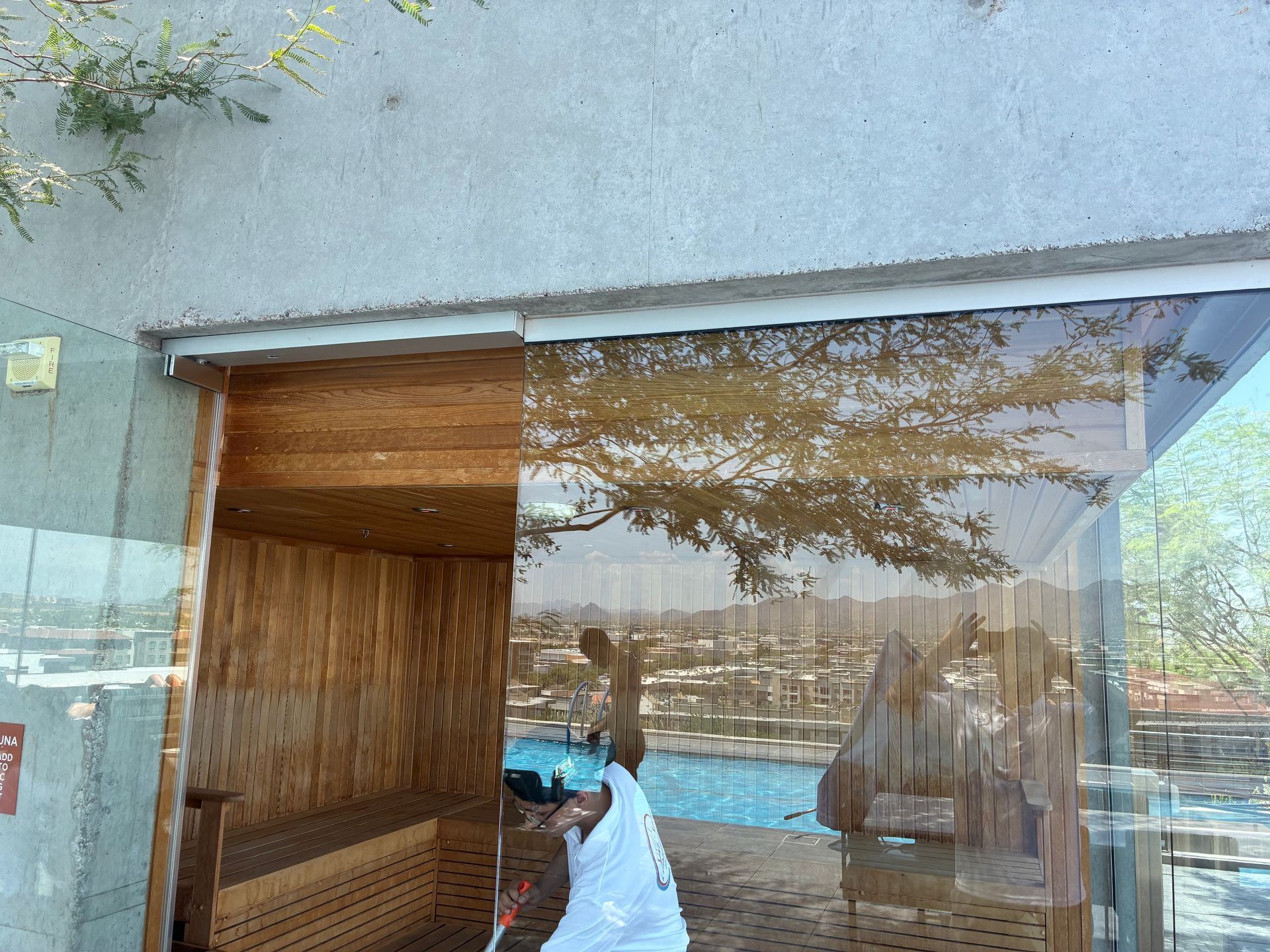 Clear plastic curtain over a wooden sauna entrance reflecting trees and a pool. A person cleans inside.