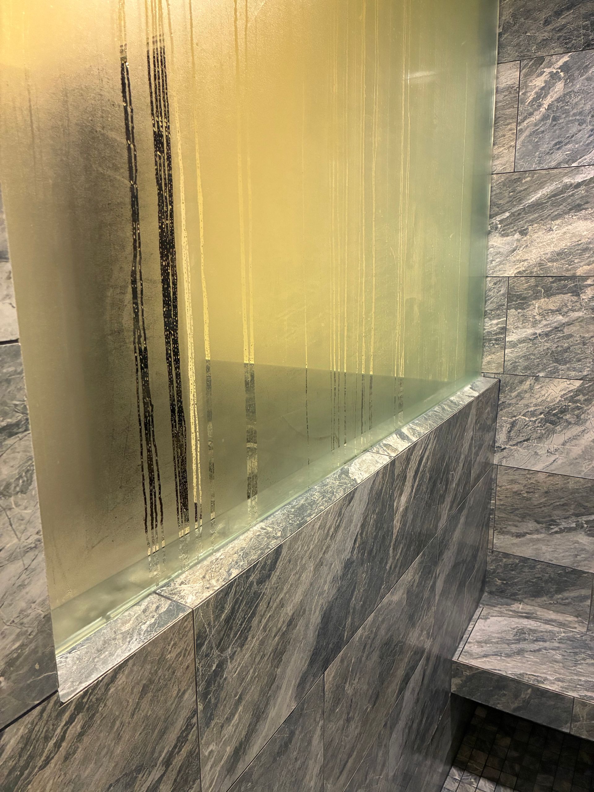 Steamy shower glass panel with vertical streaks, adjacent to a gray stone wall.