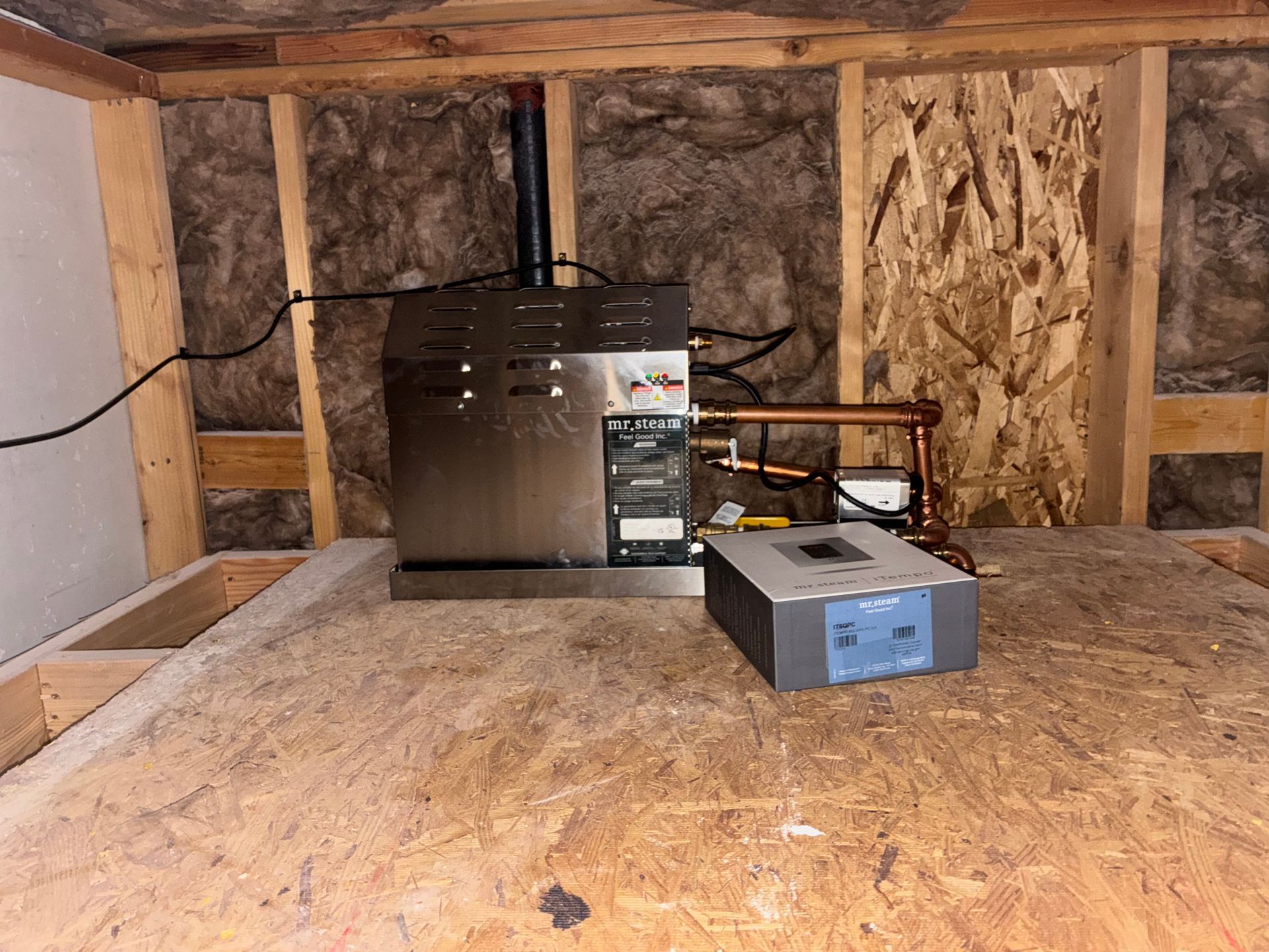 Stainless steel appliance and gray control box on a wooden platform in a basement. Copper pipes and black wires visible.