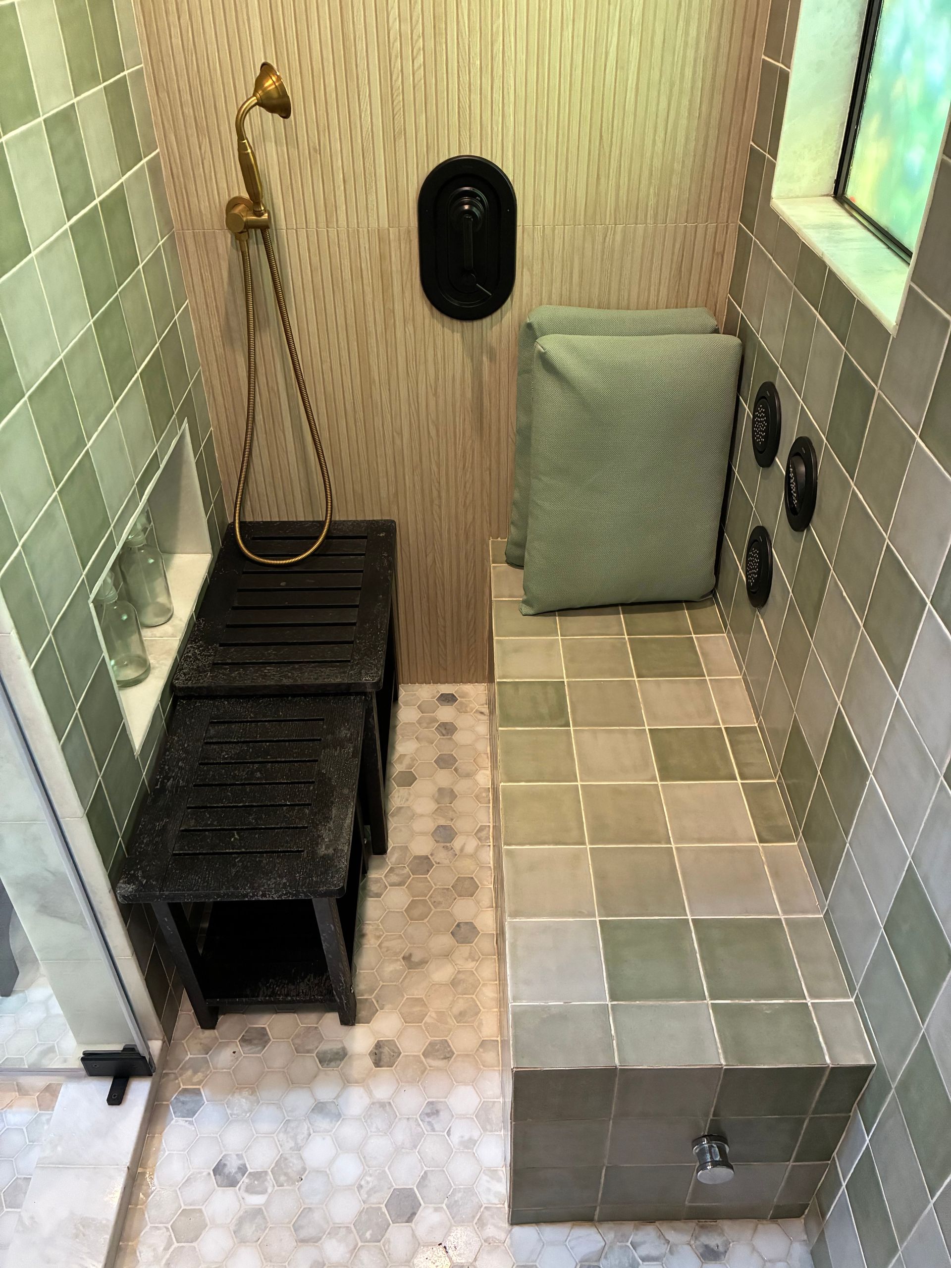 Shower with green tile, bench, black stools, and gold showerhead.