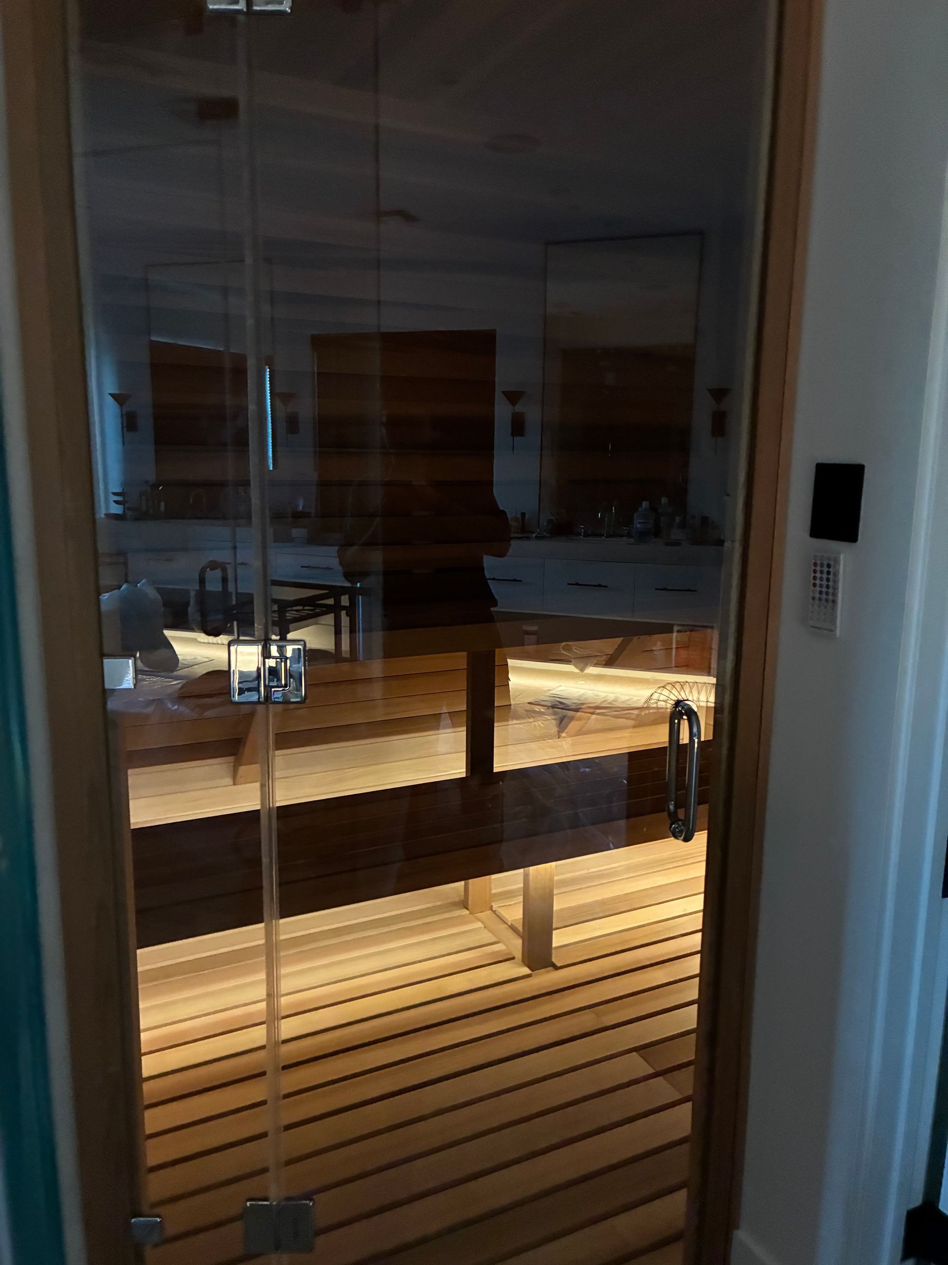 Glass-door sauna with wood interior, chrome hardware, and light wood door frame, located inside a room.