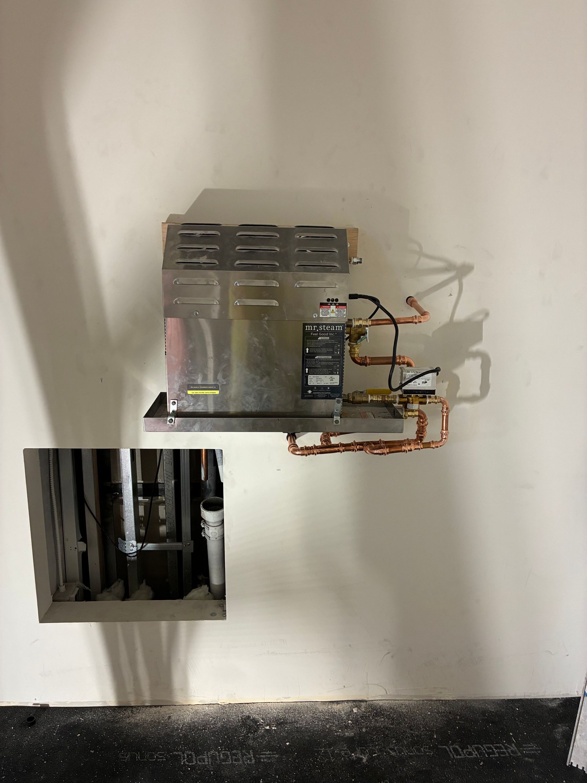 Electrical components and copper piping mounted on a wall, with a rectangular access hole below.