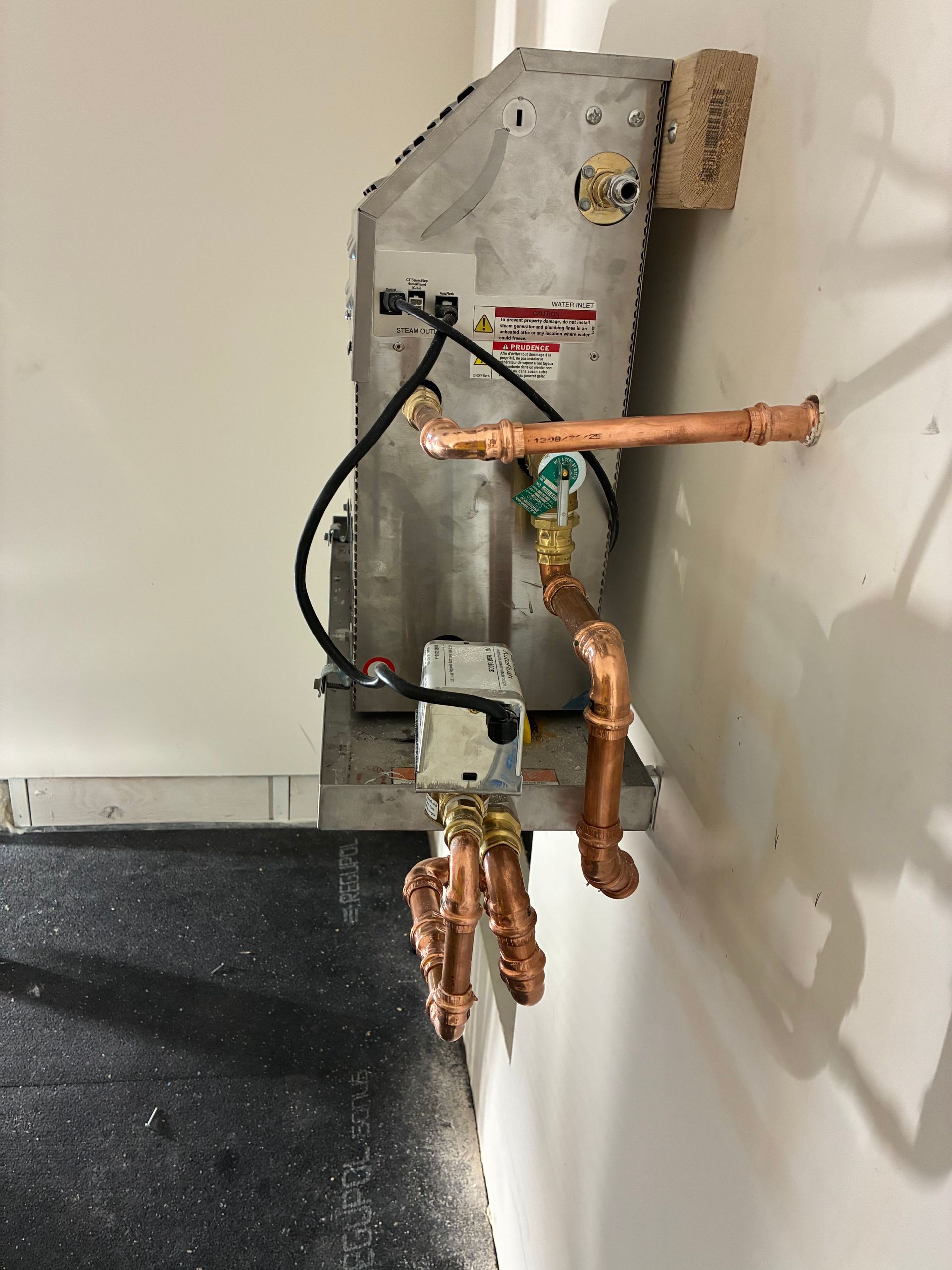 HVAC unit on a wall, with copper pipes and electrical wiring attached. Gray unit, white wall, black floor.