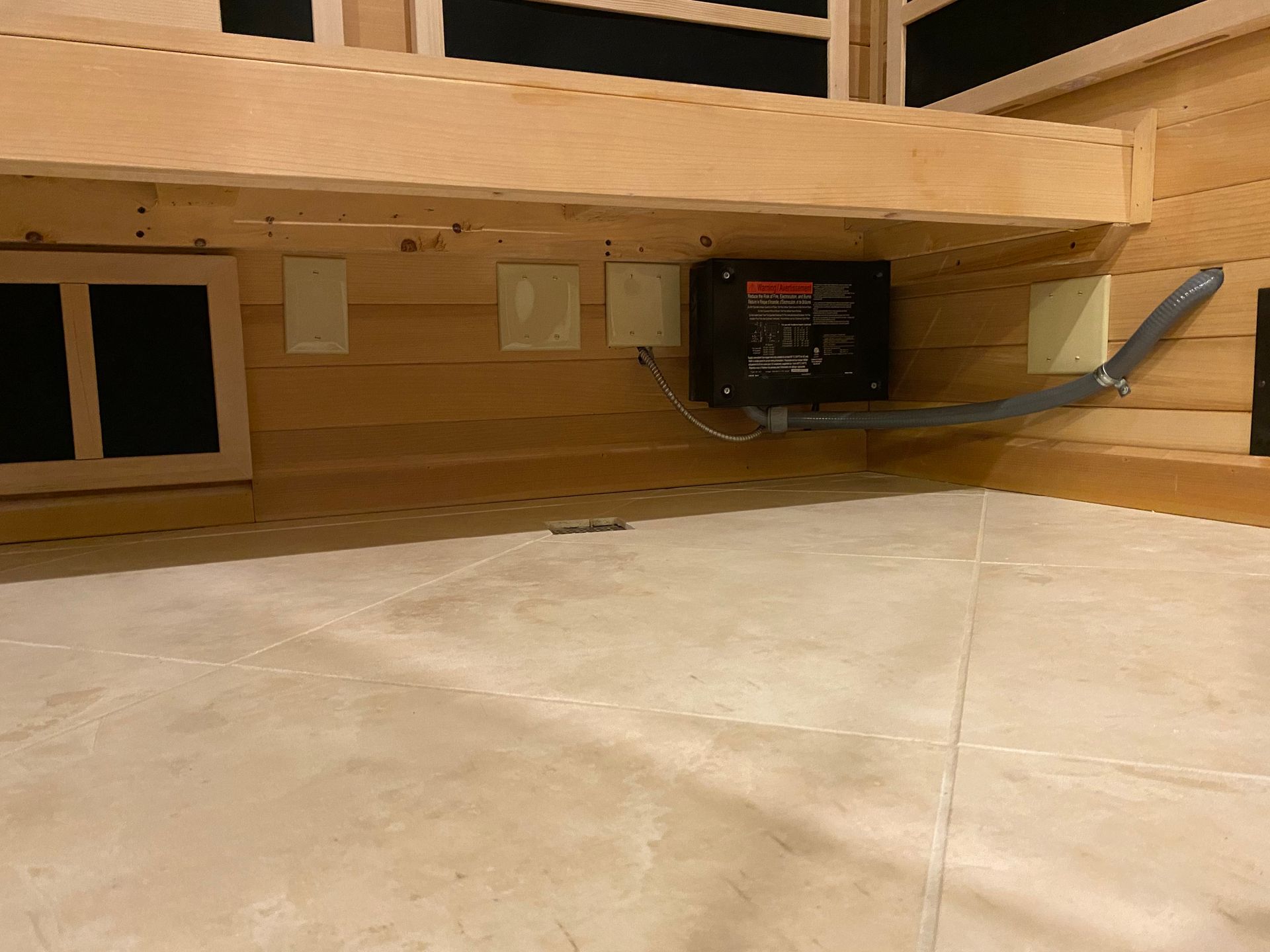 Inside of a wooden sauna with a control panel and conduit on the wall. The floor is tiled.