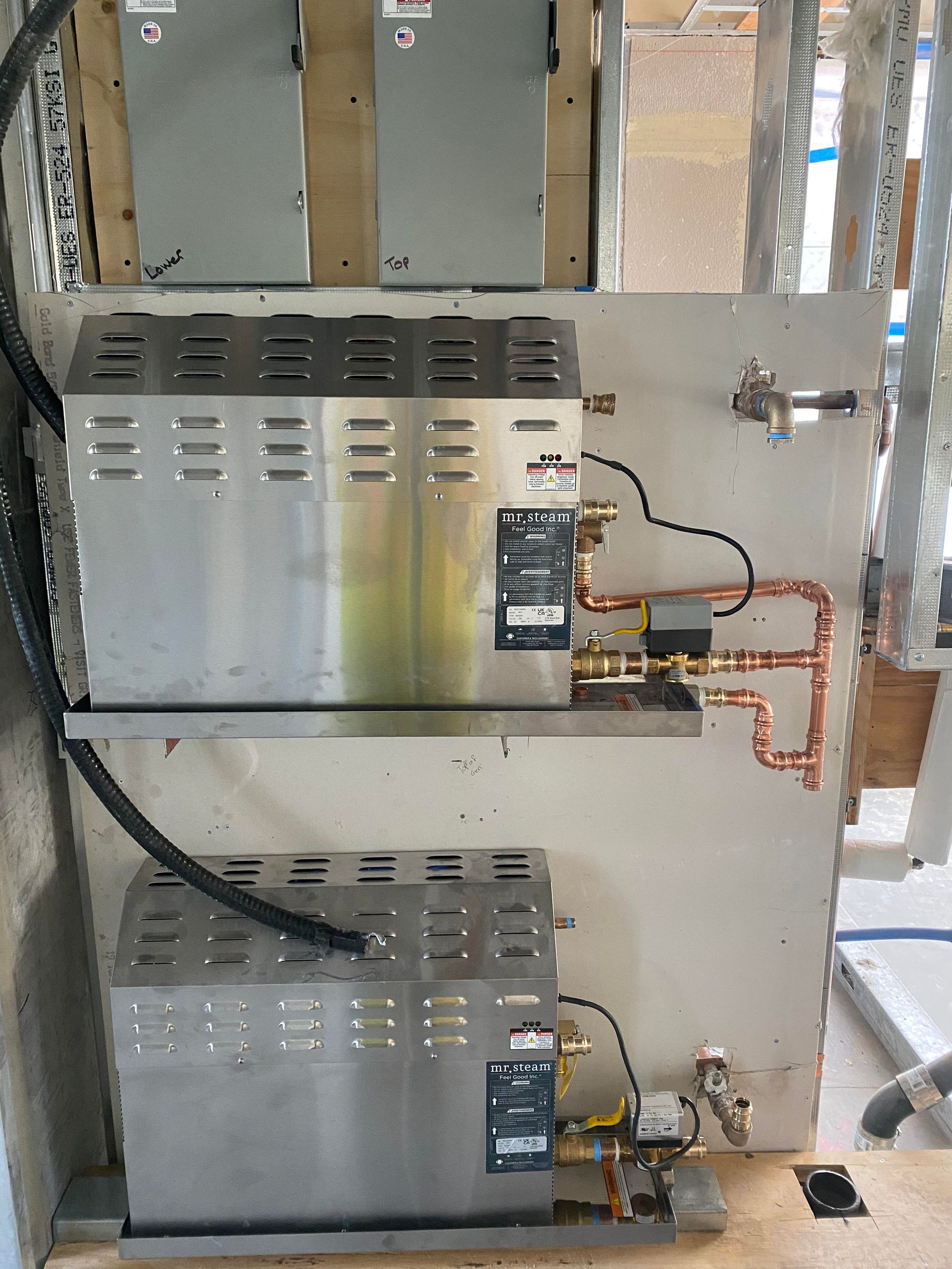 Two stainless steel steam generators mounted on a wall with copper pipes and black wiring.