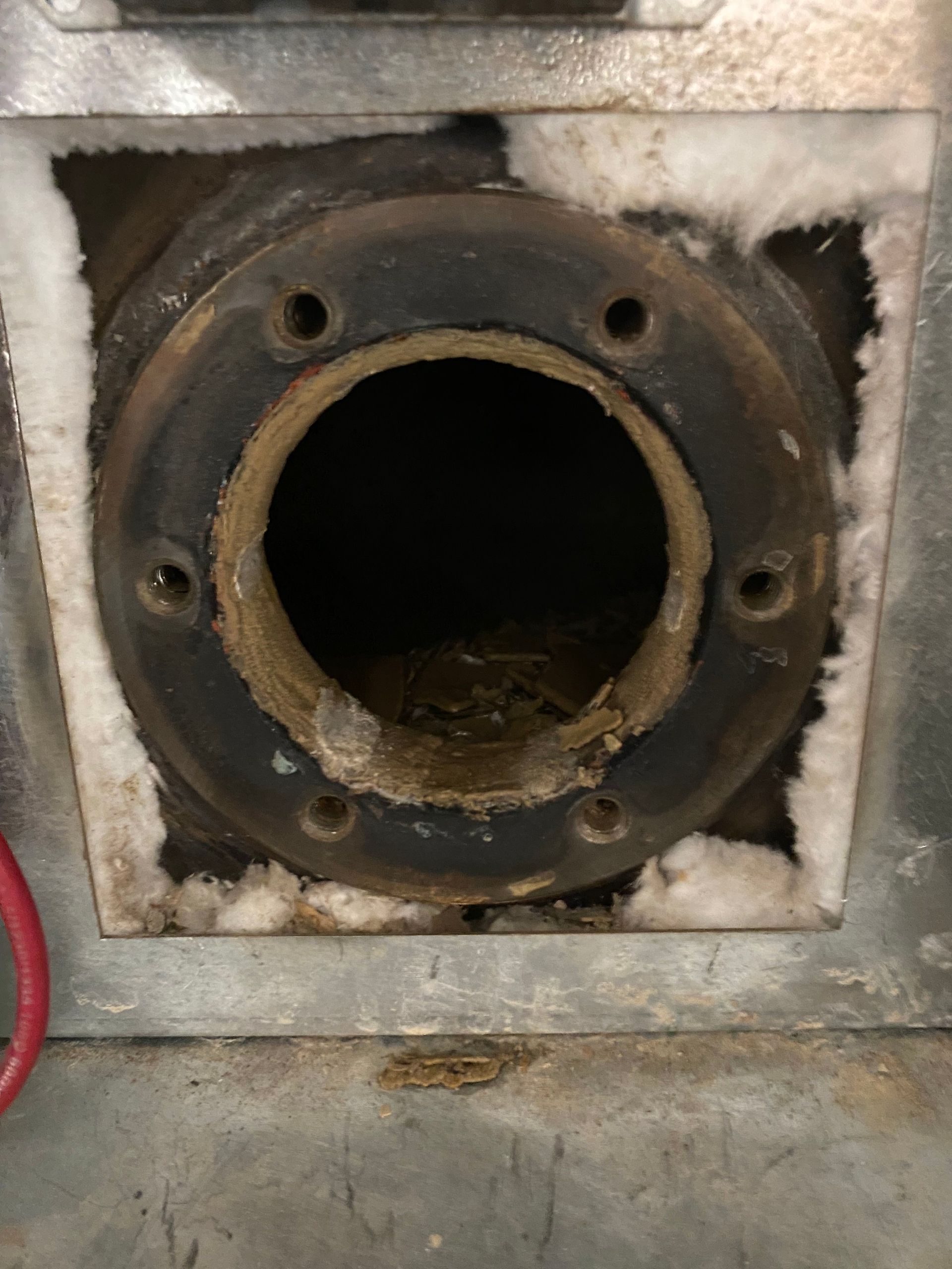 Black circular opening in a metal appliance, surrounded by insulation and bolted flange; debris inside.