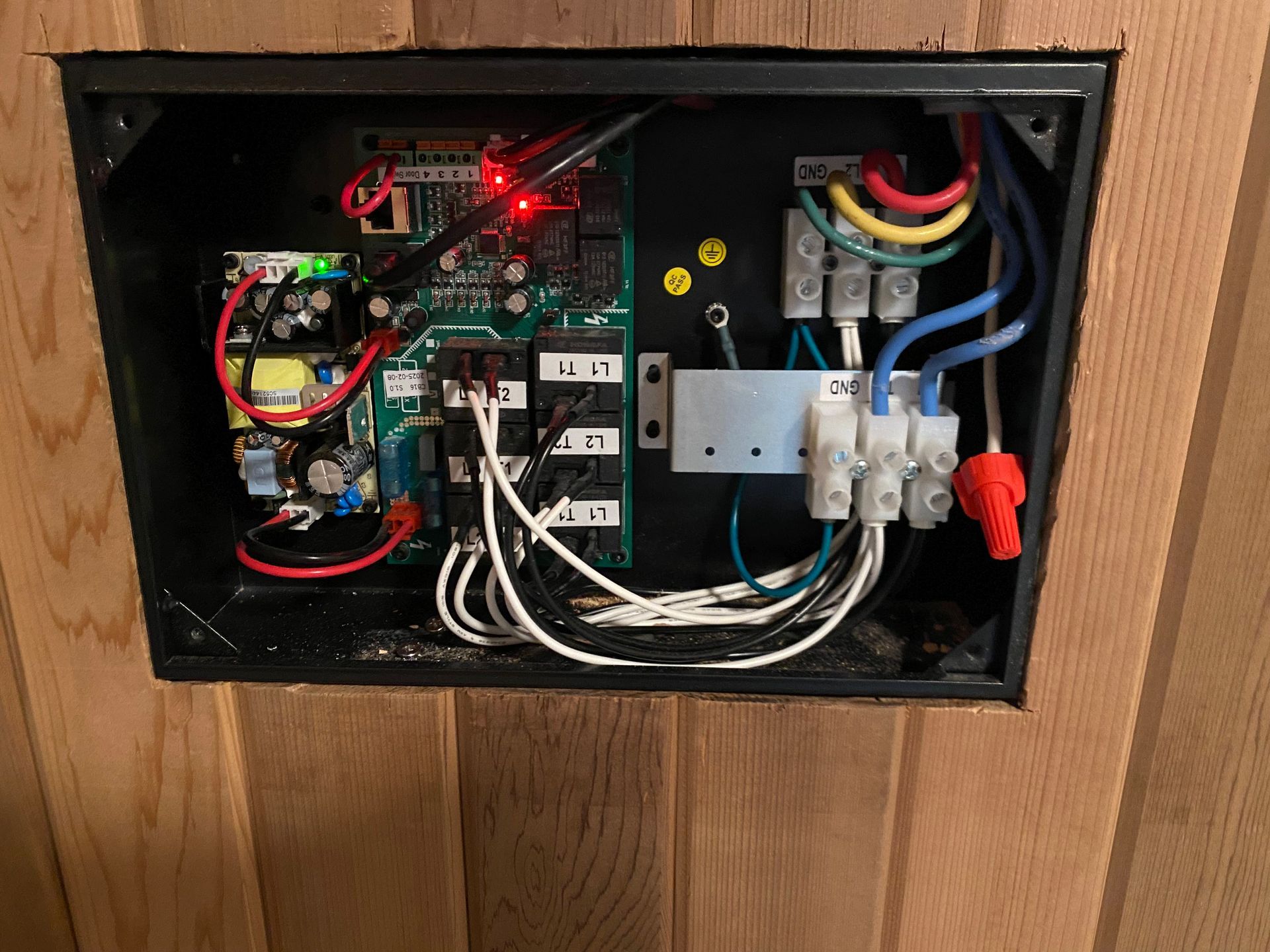 Opened electrical box on a wooden wall, containing circuit boards, wires, and connectors of various colors.