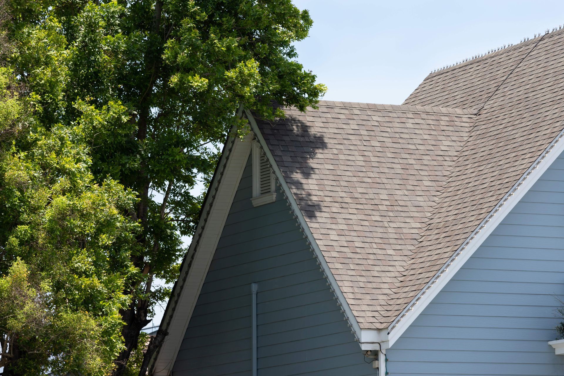 Gray shingle roof on a blue house with trees in the background under clear sky.