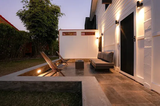 Outdoor patio with seating, lit by wall sconces, near a white building and greenery — Salisbury Concrete Contractors In Ocean Shores, NSW