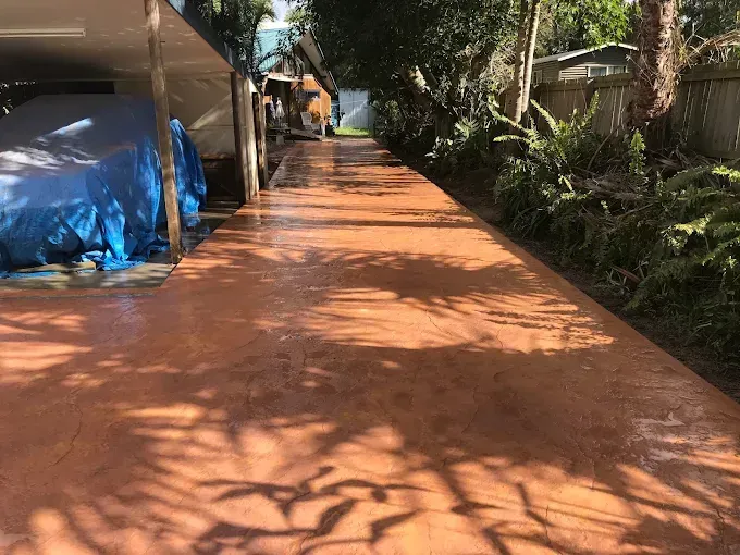 Brown, wet concrete driveway; covered car on the left, foliage on the right, trees cast shadows — Salisbury Concrete Contractors In Ocean Shores, NSW