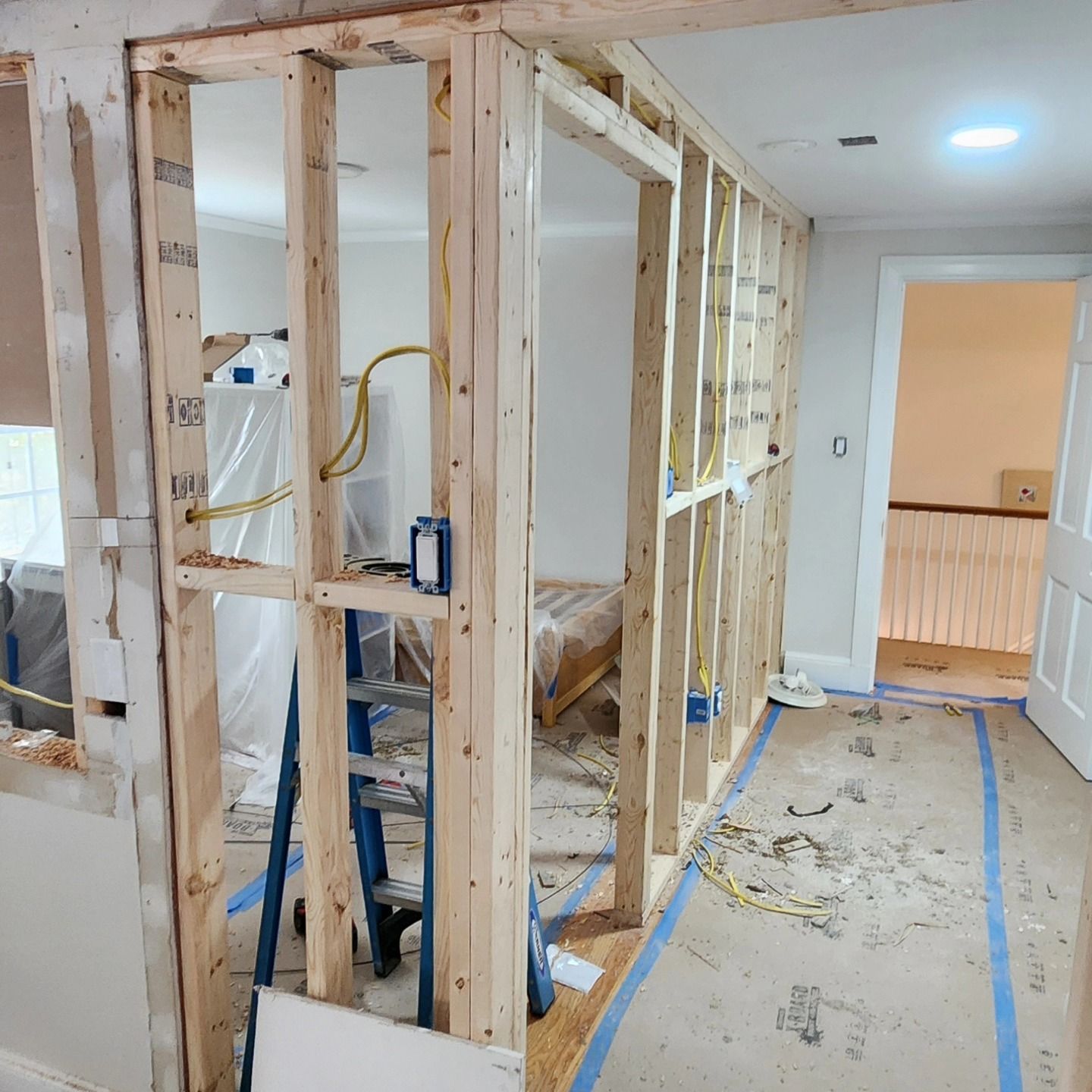 Interior room under construction, with exposed wooden studs, wiring, ladder, and a partially open doorway.