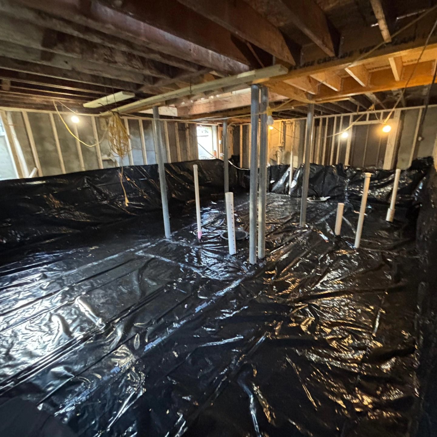 Interior view of a basement under construction, covered in black plastic sheeting. Supports and pipes are visible.