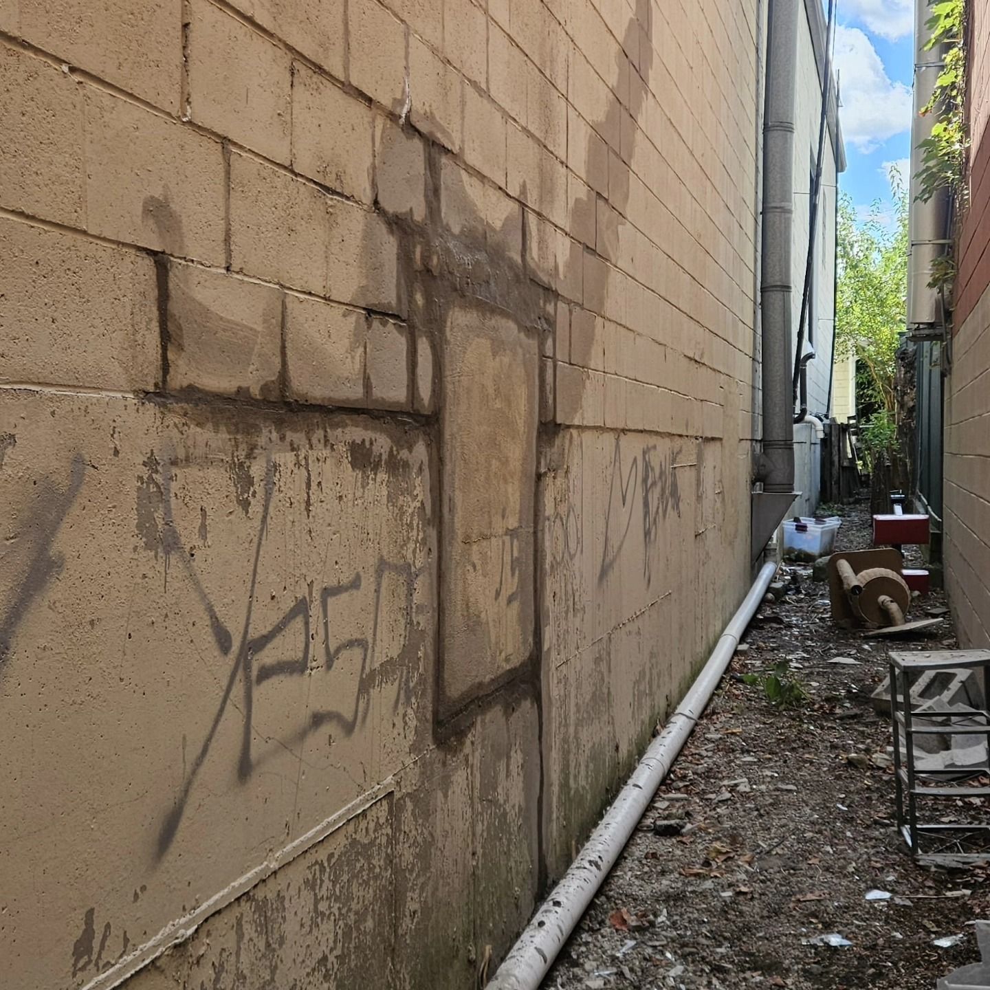 Narrow alley with spray paint on a concrete wall. A drainage pipe and debris are along the ground.