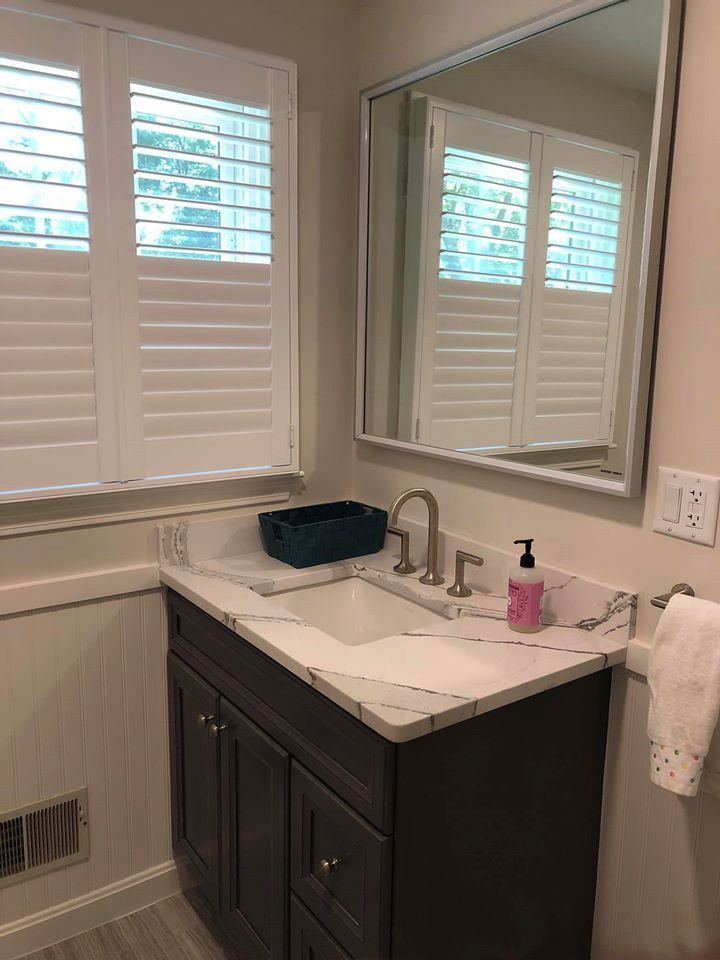 Bathroom with vanity, window with shutters, mirror. Gray vanity, white countertop, silver faucet, and a window.