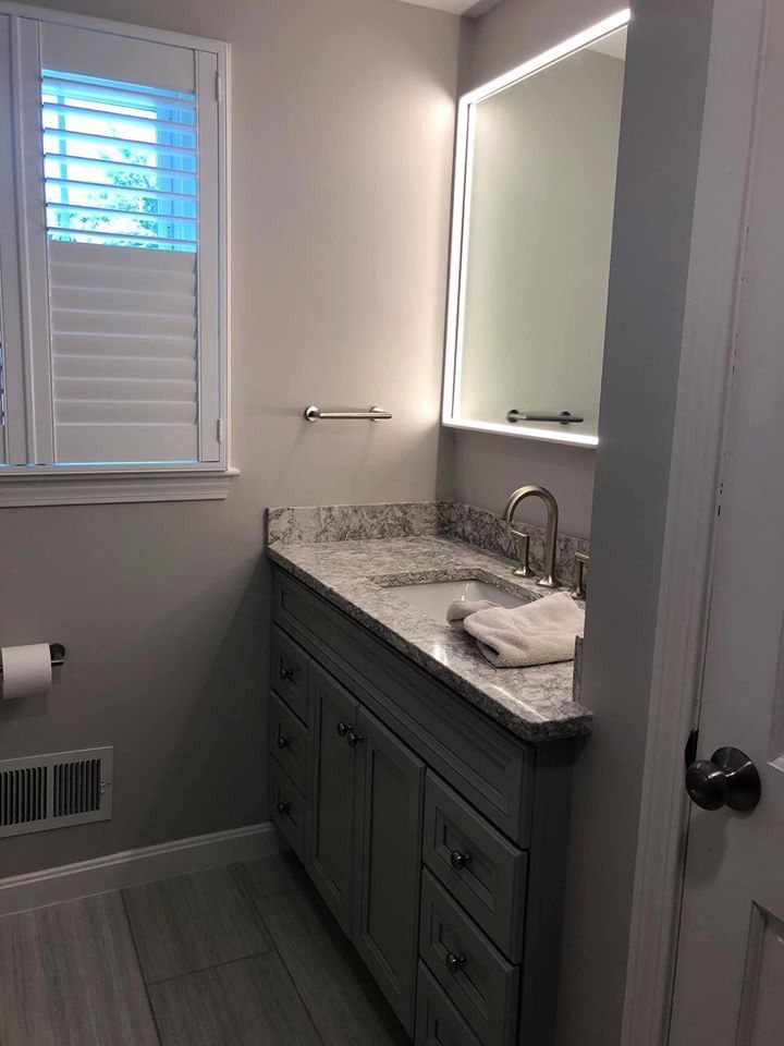 Bathroom with gray cabinets, granite countertop, mirror with lights, and window with shutters.