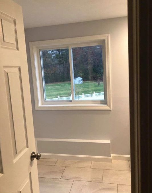 Room interior with a window overlooking a green yard. Beige floor tiles, white trim, gray walls, and a partly open white door.