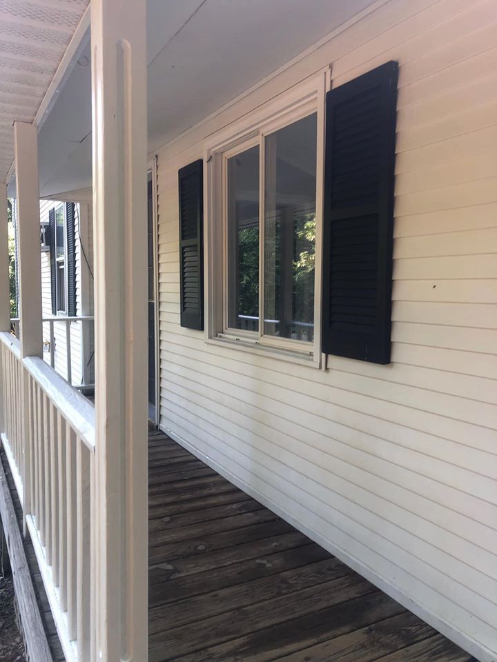 Side view of a white house porch with dark shutters, wood planks, and a railing.