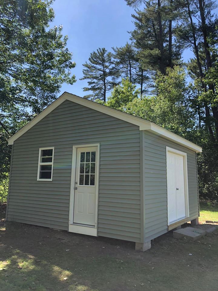 Small gray shed with white door and window, standing in a yard surrounded by trees.