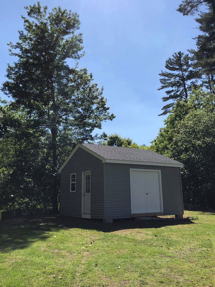 Blue-gray shed with a white door and garage door, surrounded by trees on a sunny day.