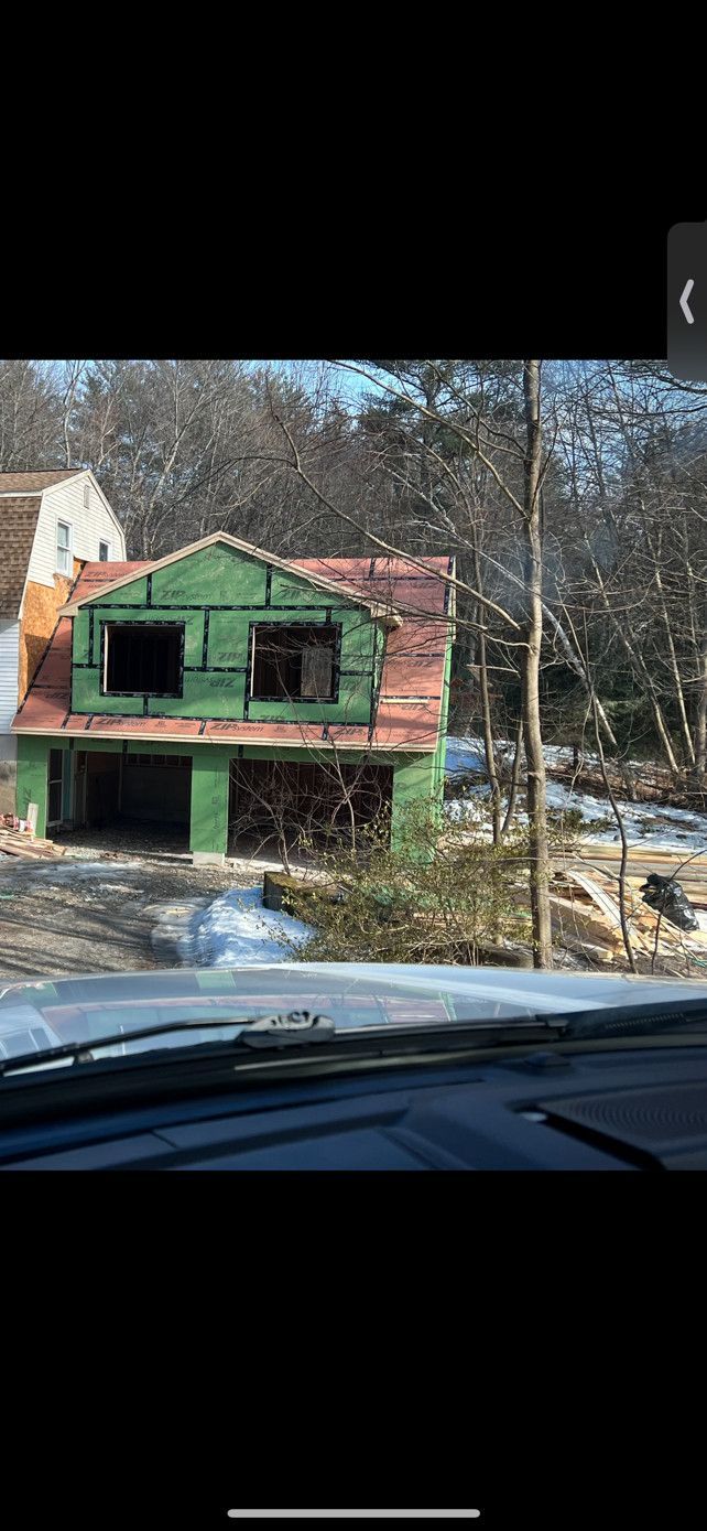 A two-story house under construction, green siding, brown roof, surrounded by trees, viewed from a vehicle.