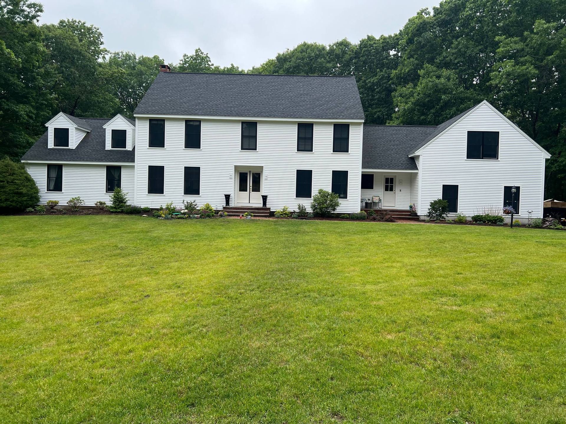 White two-story house with black shutters and a dark roof, set in a green yard surrounded by trees.