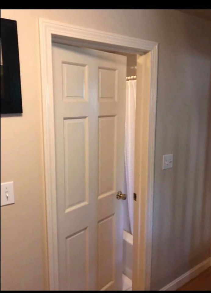White six-panel door slightly ajar, revealing a shower curtain and bathroom. Beige walls and trim.