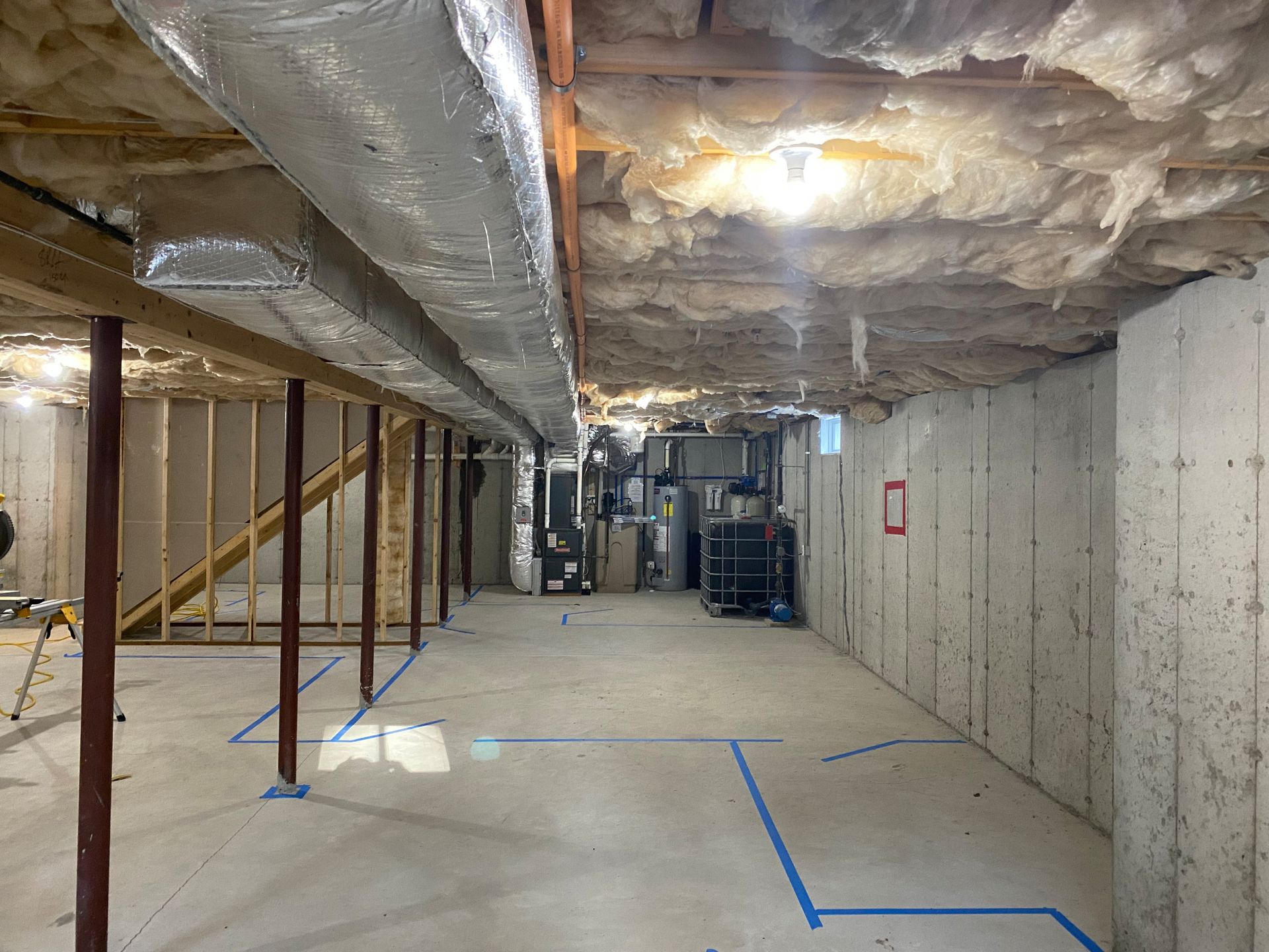 Unfinished basement with ductwork, concrete walls, wooden beams, and blue tape markings on the floor.