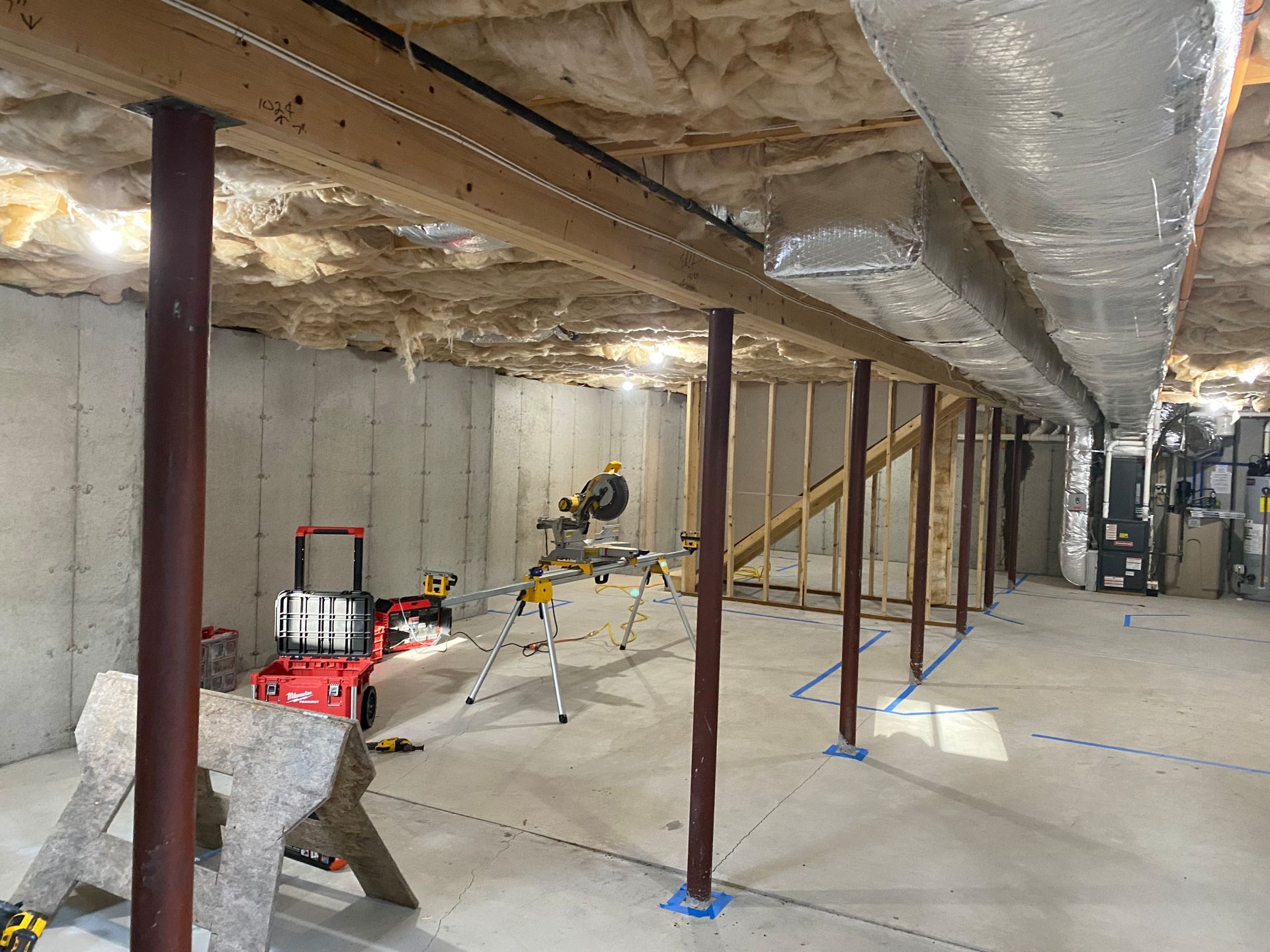 Basement under construction with saw, tools, wooden beams, support columns, and concrete walls.
