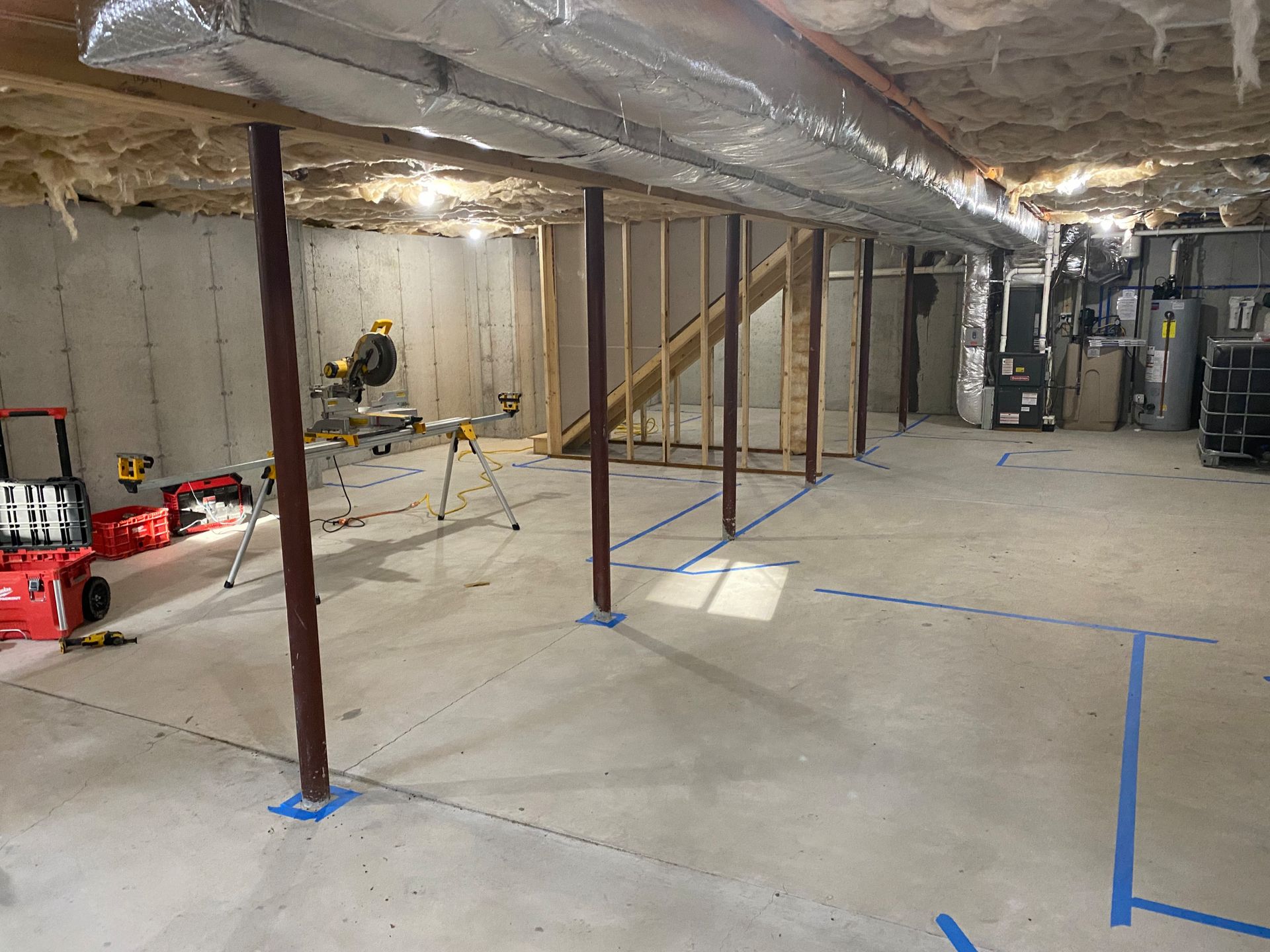 Basement under construction. Concrete floor, support beams, framing, insulation. Blue tape marks out areas.