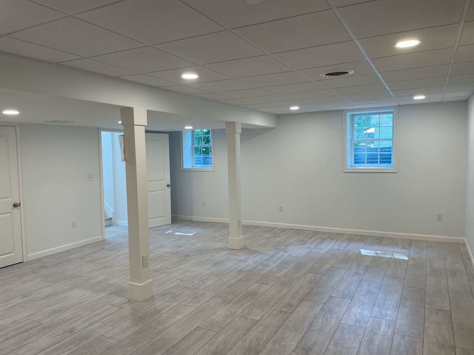 Empty, newly-renovated basement with gray flooring, white walls, support beams, recessed lighting, and windows.