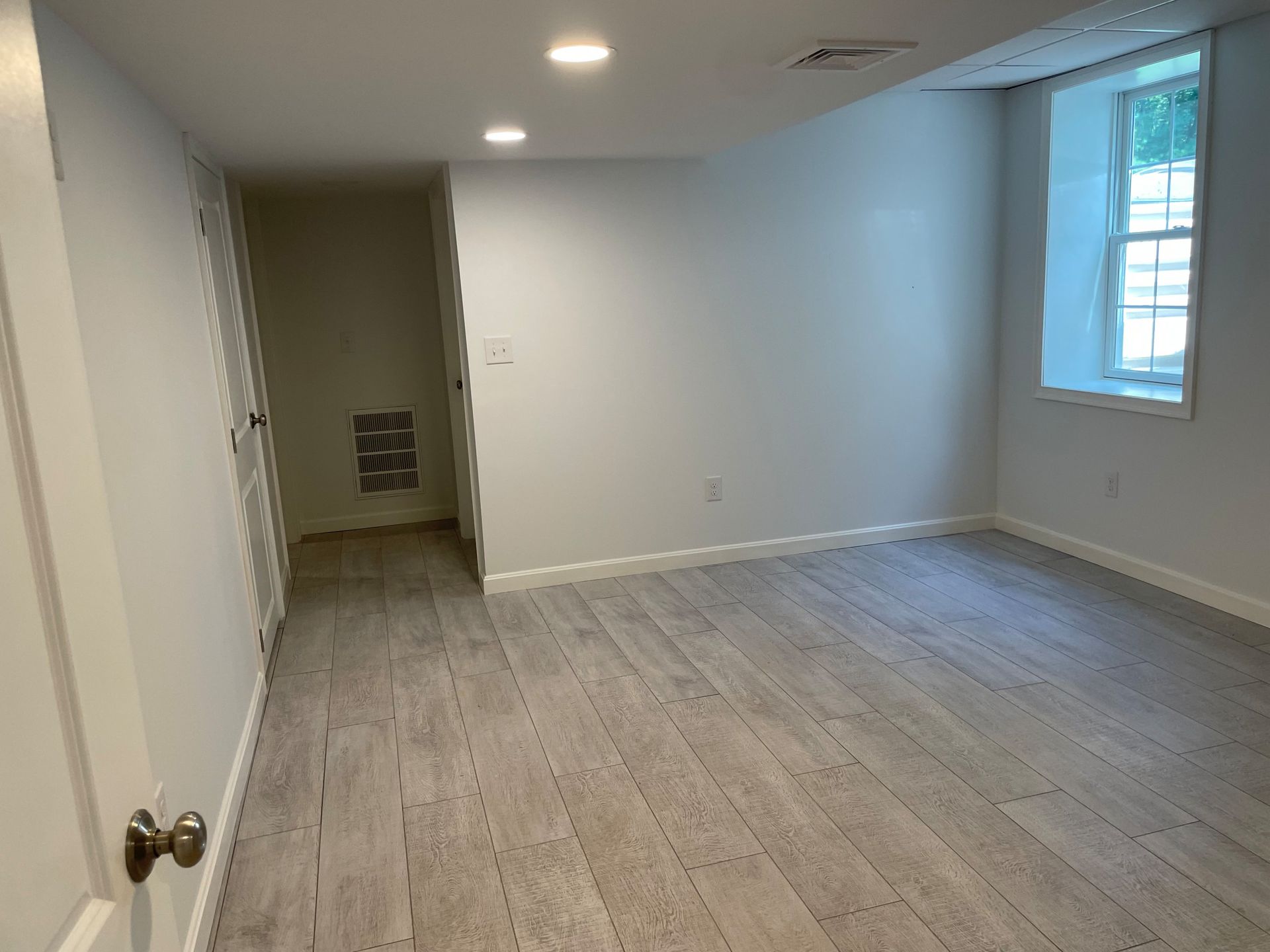 Empty room with light gray wood-look flooring, white walls, and a small window with sunlight.